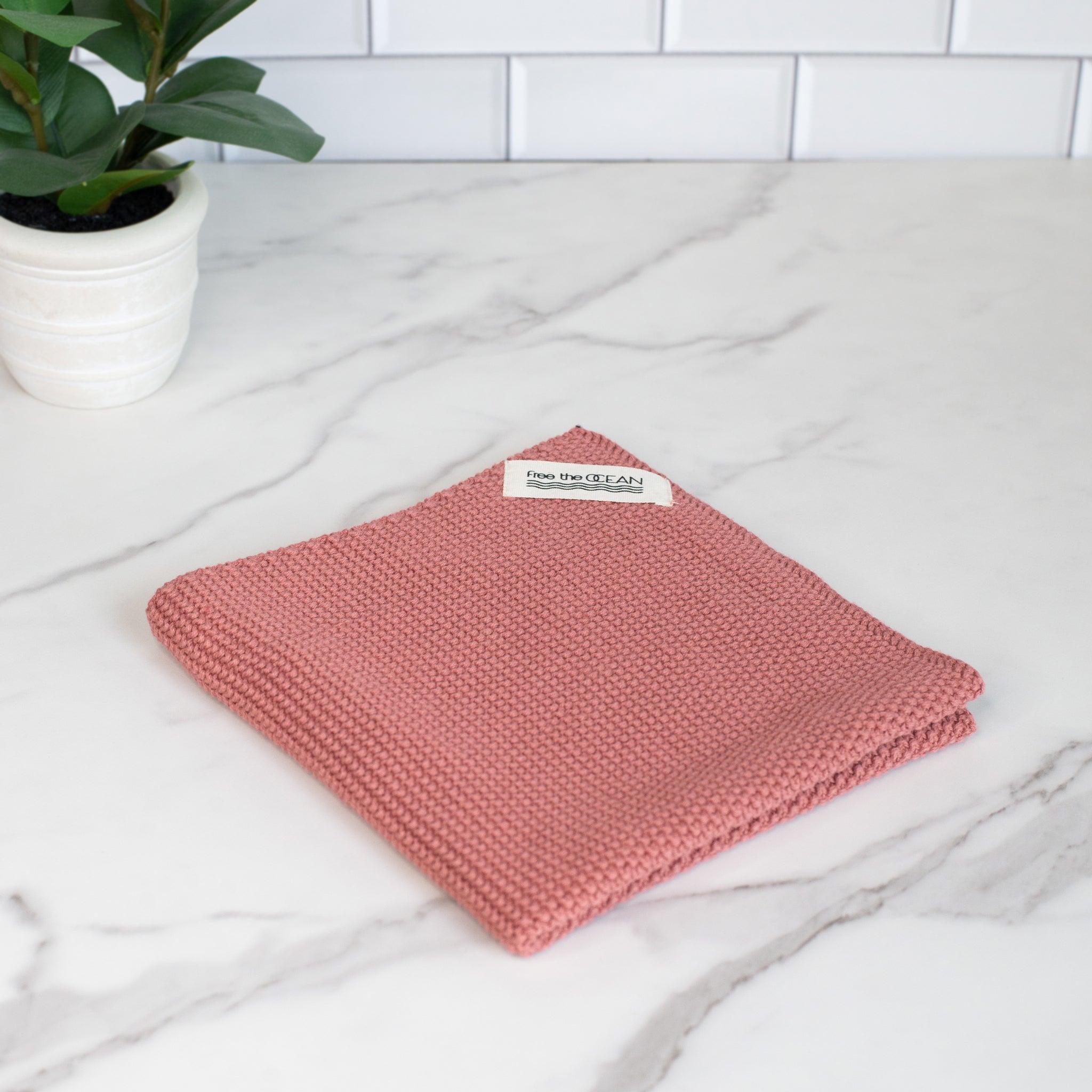 A square tightly-woven cleaning cloth in a muted rose color with a small white tag in one corner that says "free the OCEAN" in black lettering.