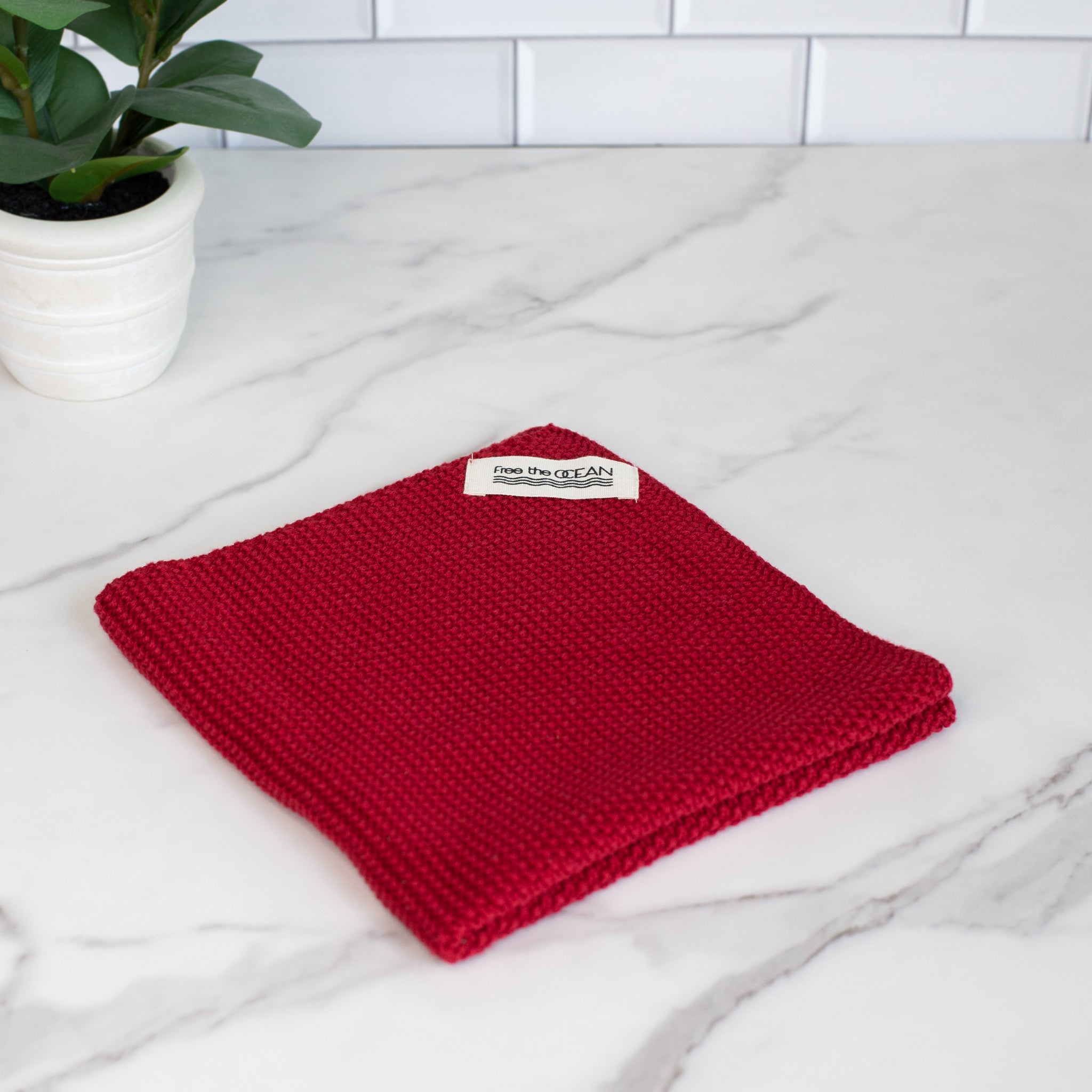 A square cleaning cloth in a rich dark red color featuring a waffle-like texture. A small white tag is visible in one corner displaying the words "free the OCEAN".