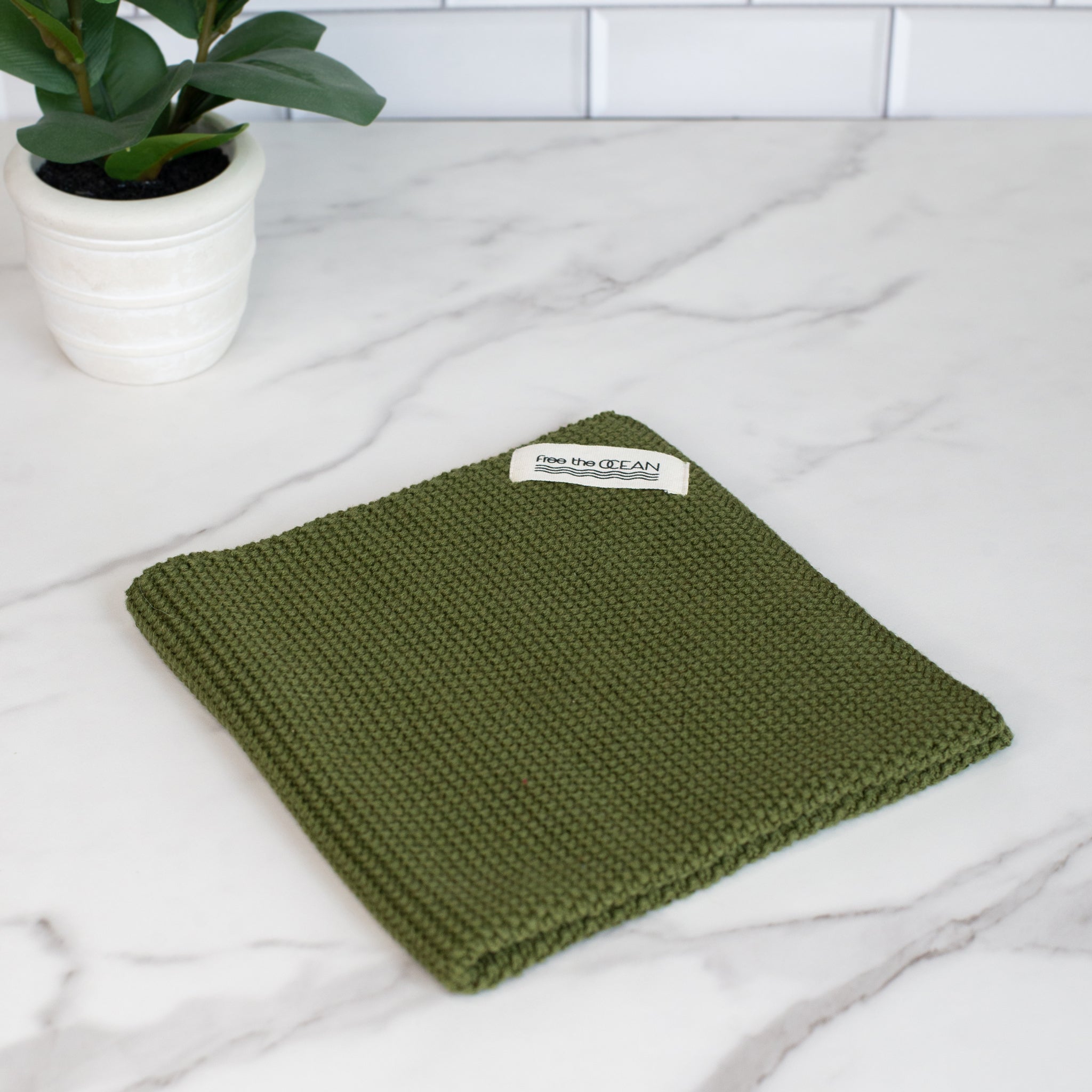 A square cleaning cloth with a waffle texture in a deep olive green color. It has a small white tag in one corner with "free the OCEAN" written in black.