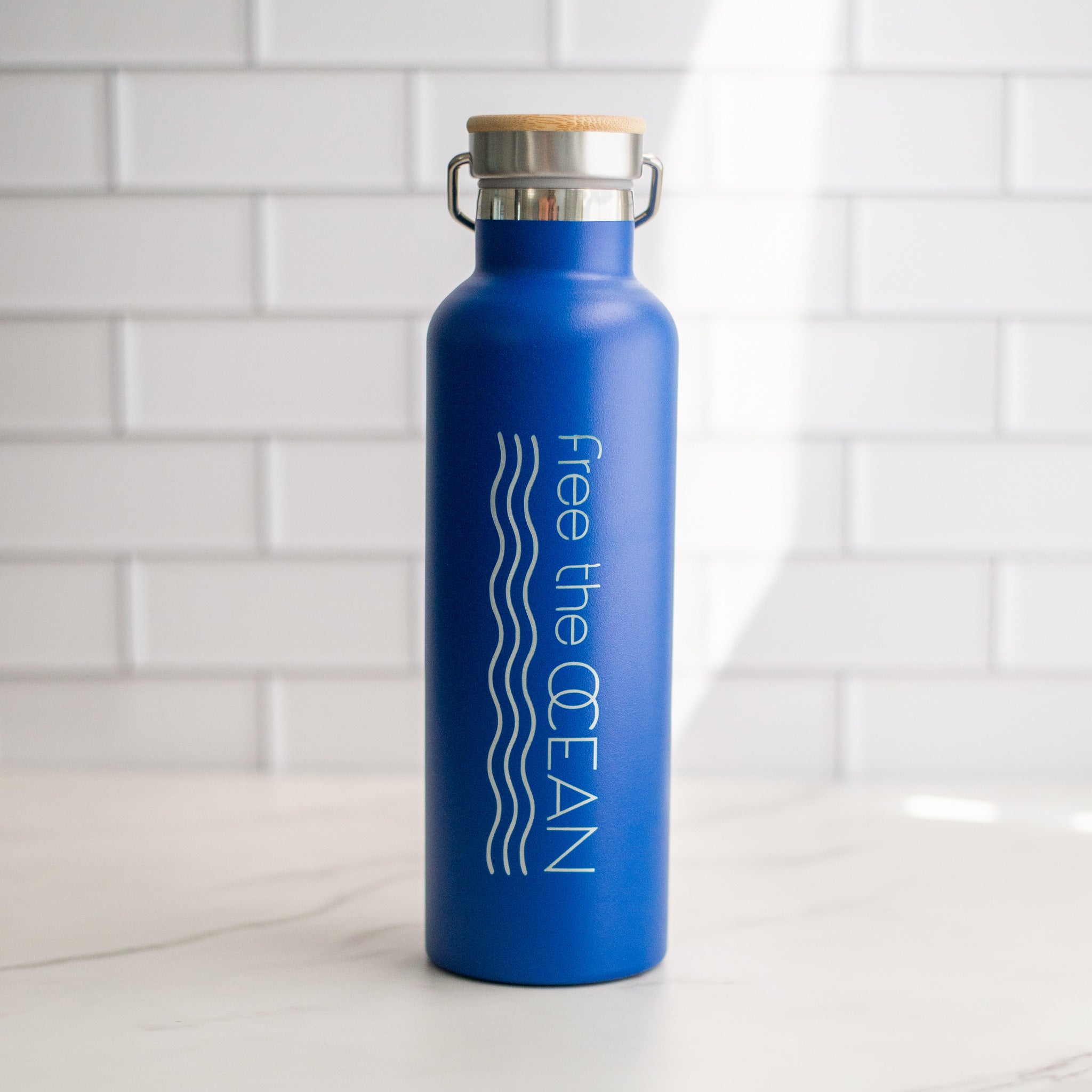 A cobalt blue reusable water bottle with a bamboo lid and a metal handle. The words "Free the OCEAN" and two wavy lines in white are visible.