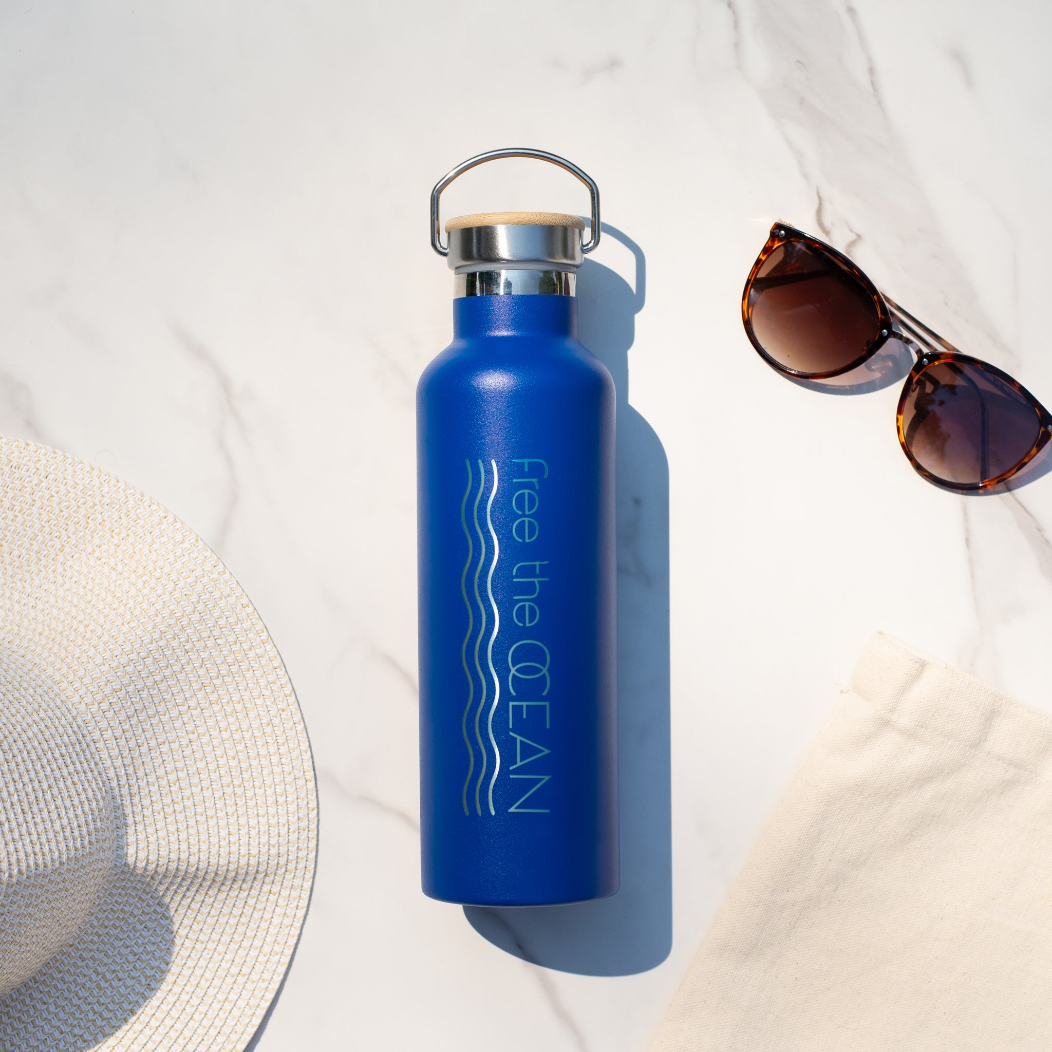 A blue reusable water bottle with a bamboo lid and a metal handle. It is lying on a marble surface next to a white sun hat and a pair of sunglasses.