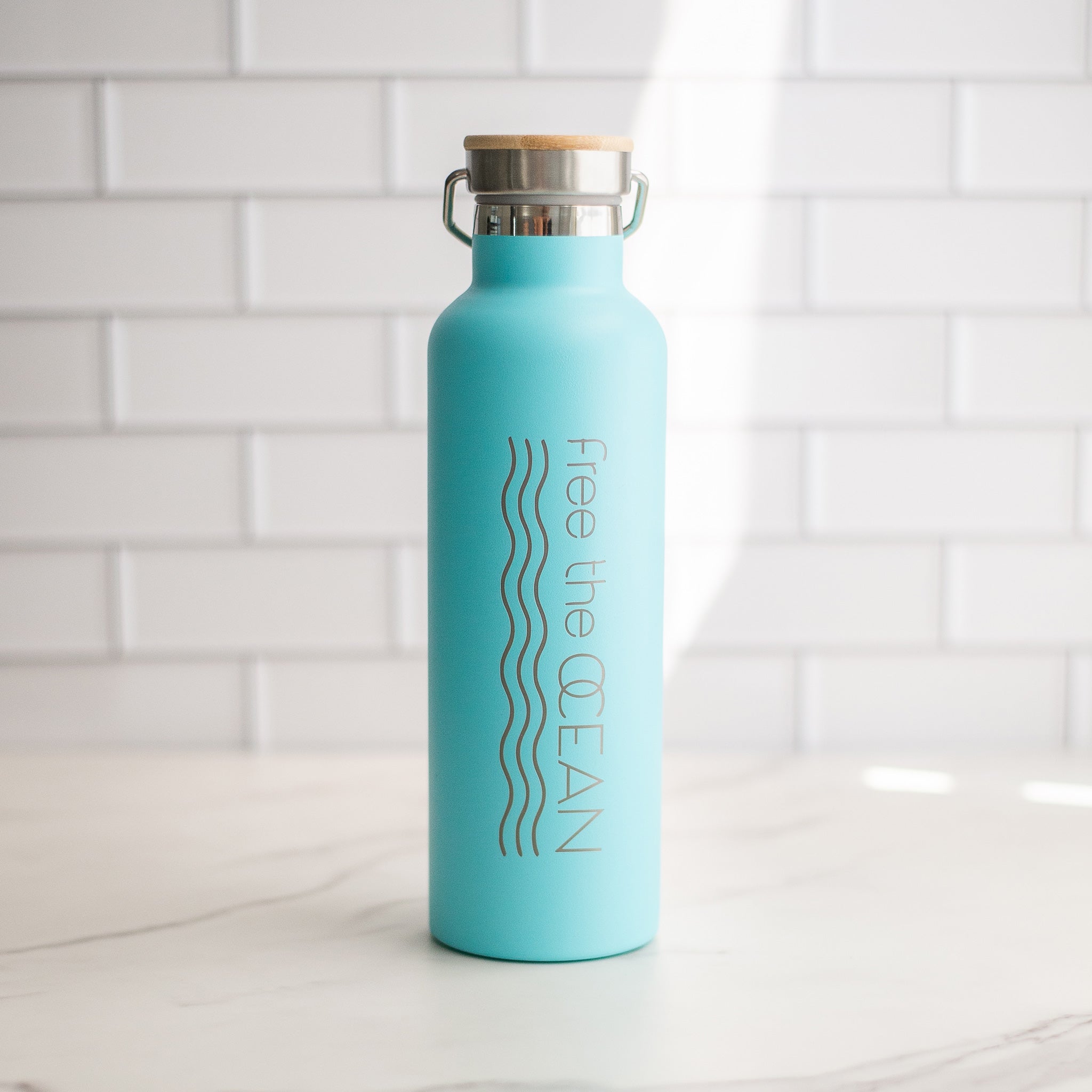 A turquoise reusable water bottle with a bamboo lid and a metal handle is on a white marble counter in front of white tiled walls.