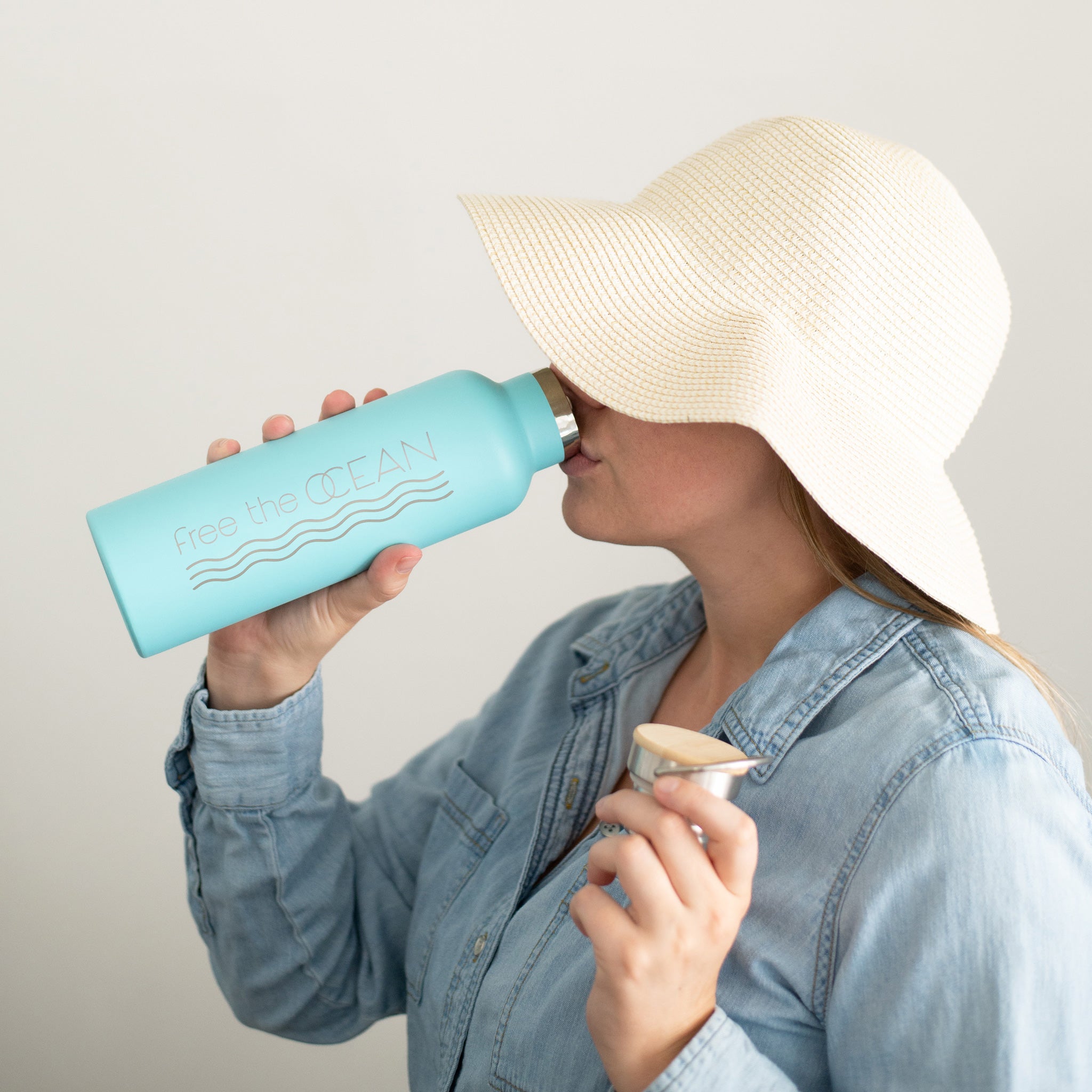 A person wearing a wide-brimmed straw hat and a denim shirt drinks from a light blue metal water bottle. The bottle has the words "free the OCEAN" printed on it. The user is enjoying a sunny day, staying hydrated with their sustainable water bottle.