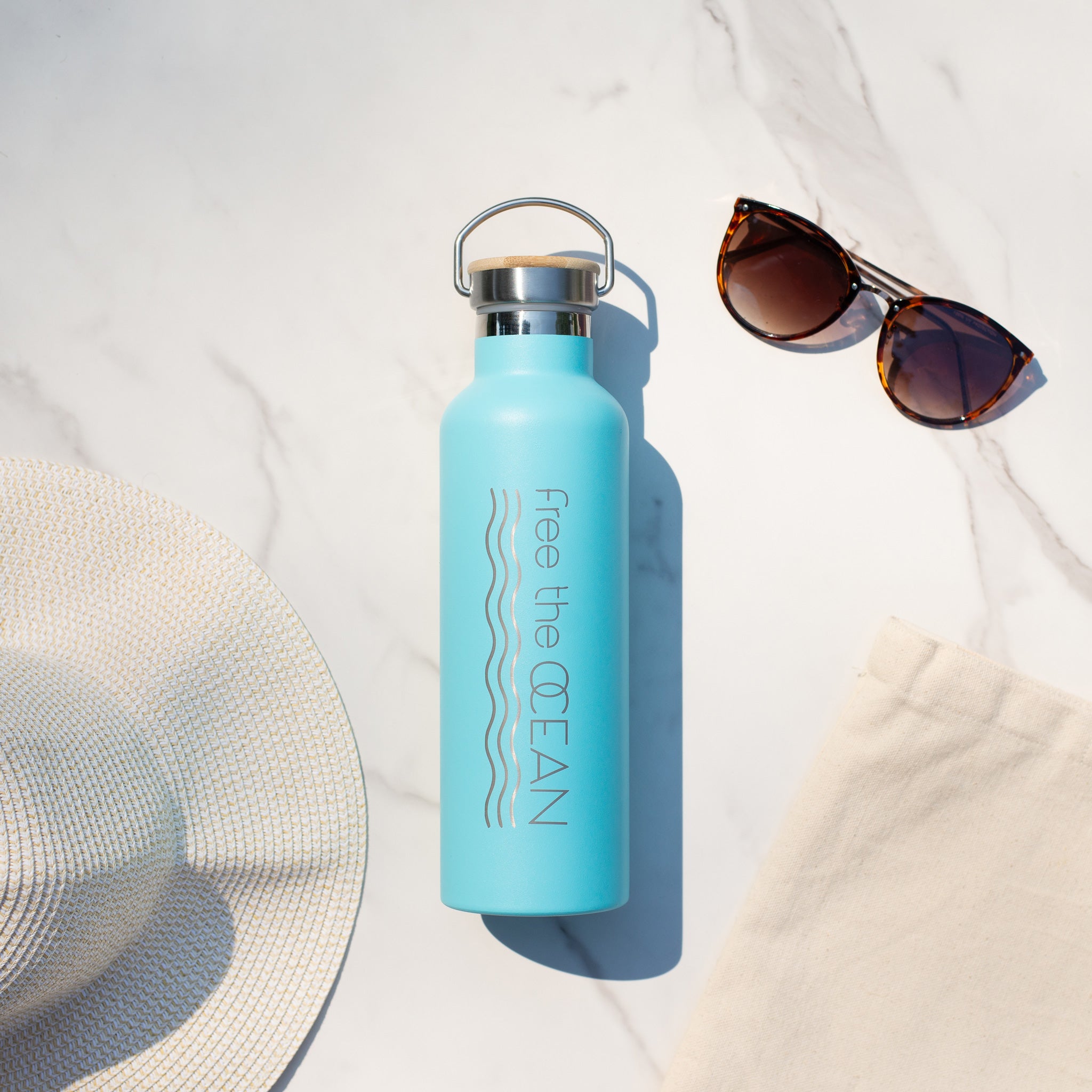 A turquoise reusable water bottle with a bamboo lid and a metal handle is on a marble surface next to a white sun hat and a pair of sunglasses.