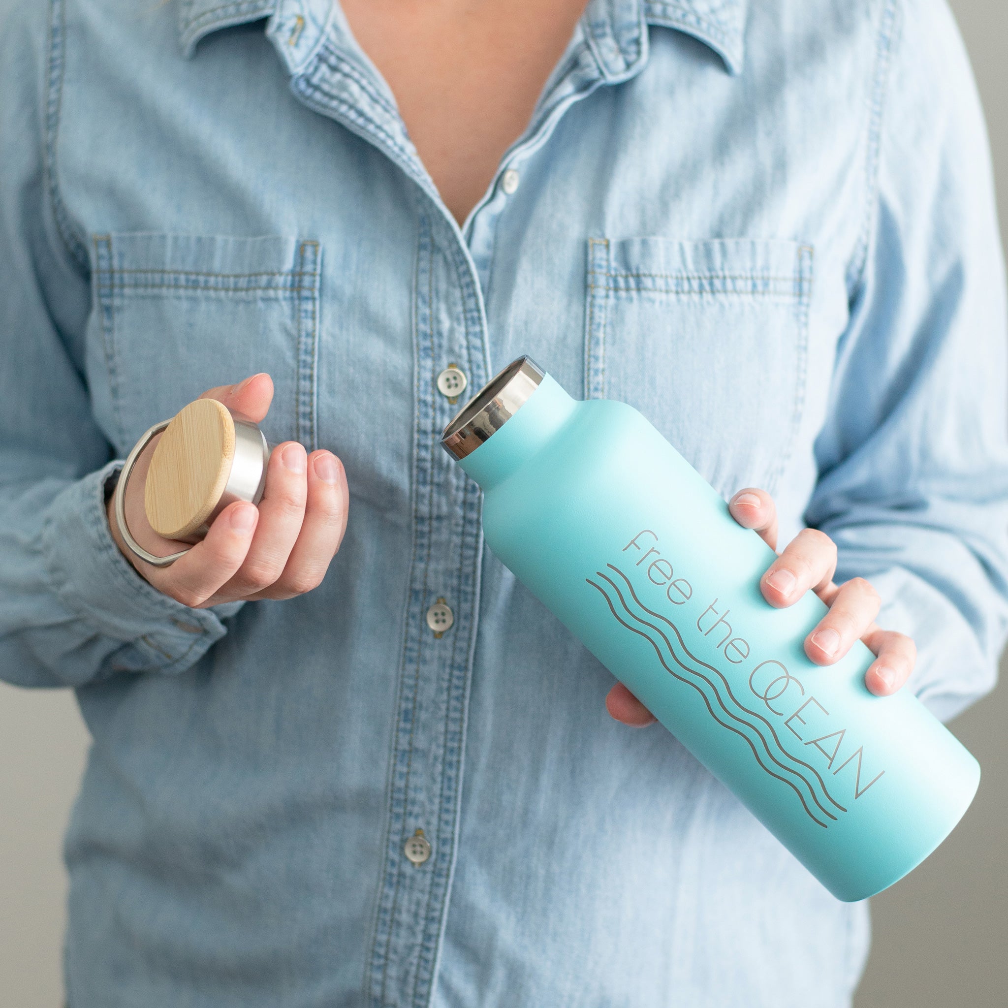 A person wearing a denim shirt holds a turquoise water bottle. The person holds the lid with its bamboo top in one hand and the bottle in the other.