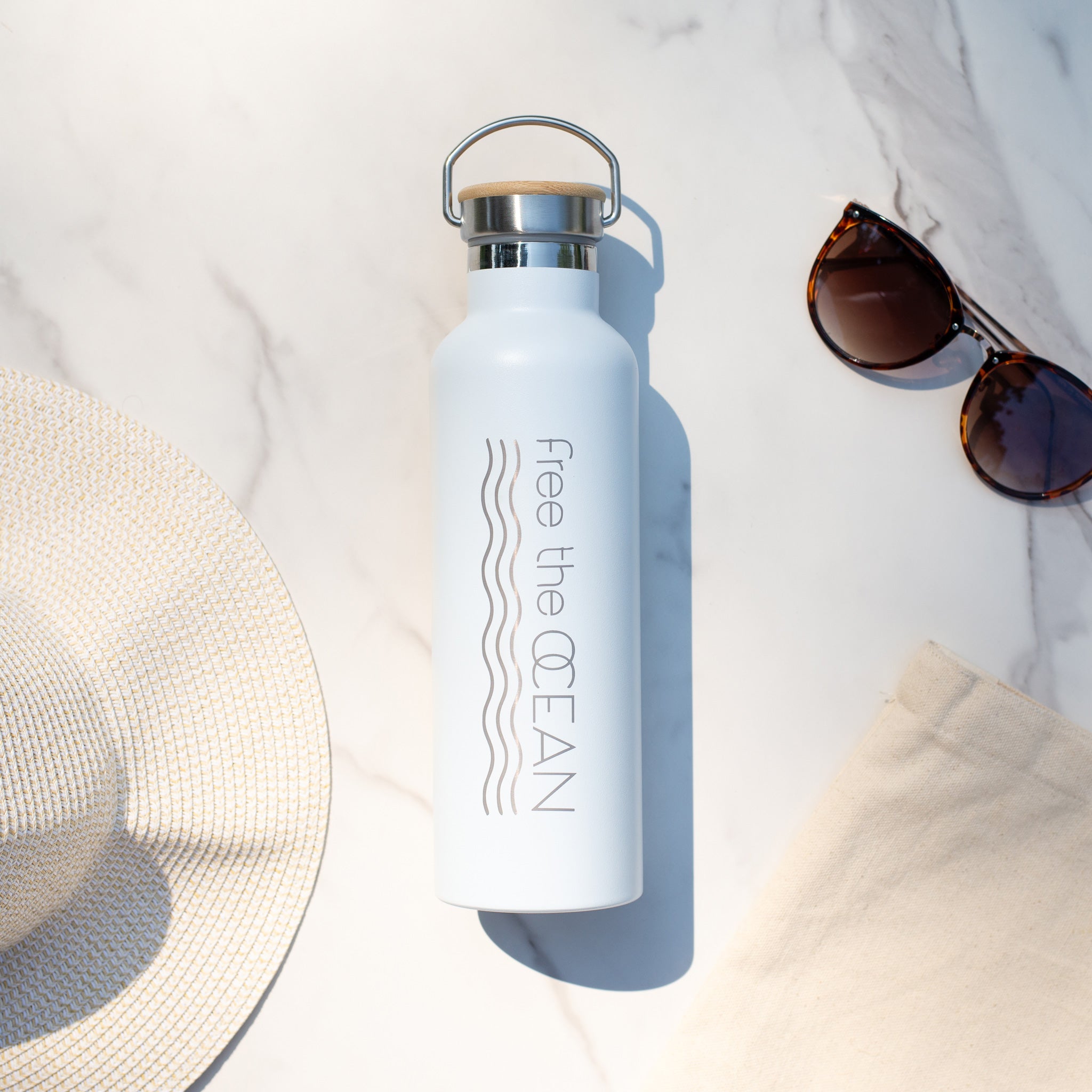 A white reusable water bottle with a bamboo lid and a metal handle. It has the words "Free the OCEAN" and two wavy lines in gray on the side. A straw hat and sunglasses are nearby.