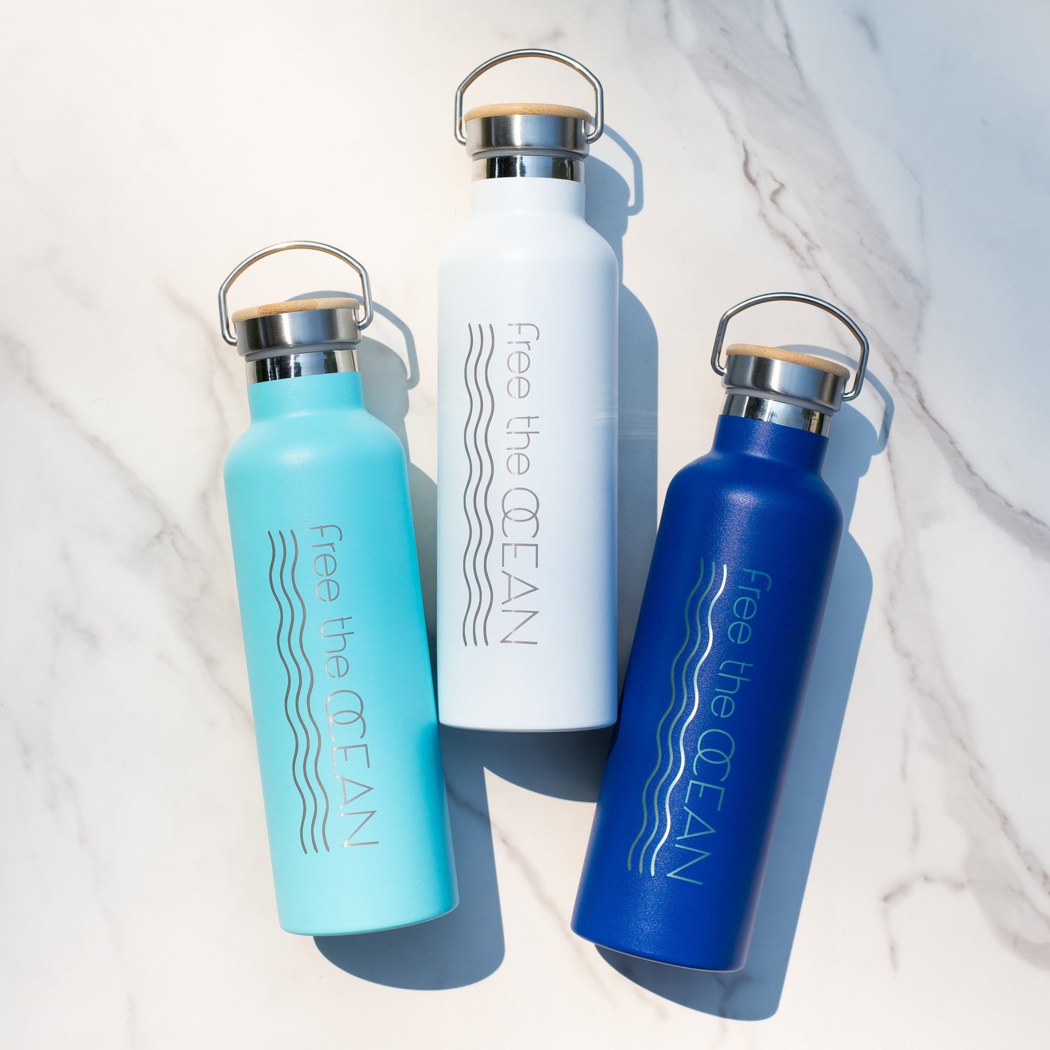 The image shows three water bottles in a diagonal line on a marble surface. The colors are turquoise white and blue. They all have bamboo lids and metal handles.