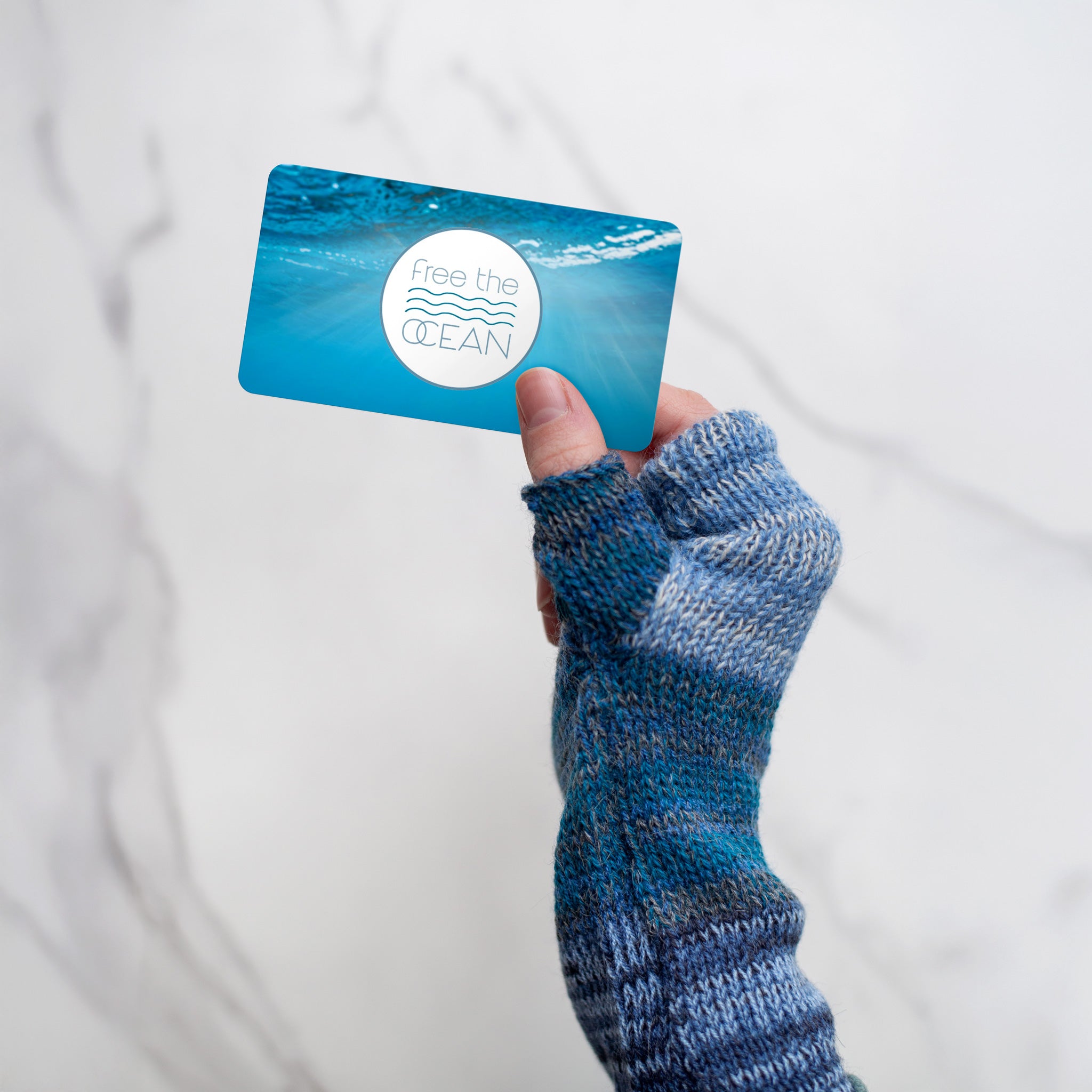 Close-up of a hand wearing a blue fingerless mitten holding a Free the Ocean gift card, highlighting the perfect eco-conscious present for friends and family.