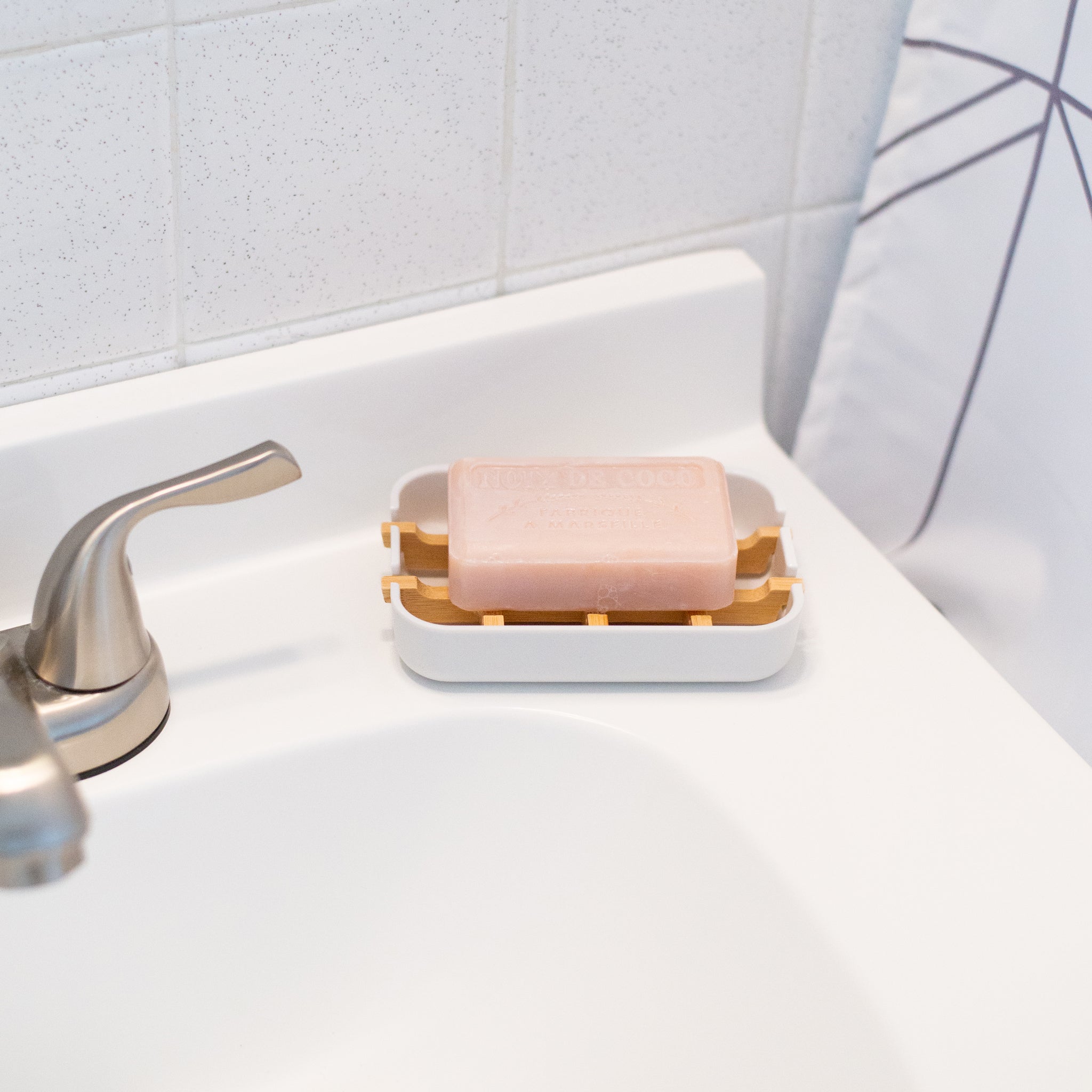 A coconut French soap bar sits on a white dish with bamboo slats, waiting to be used on a clean bathroom counter.