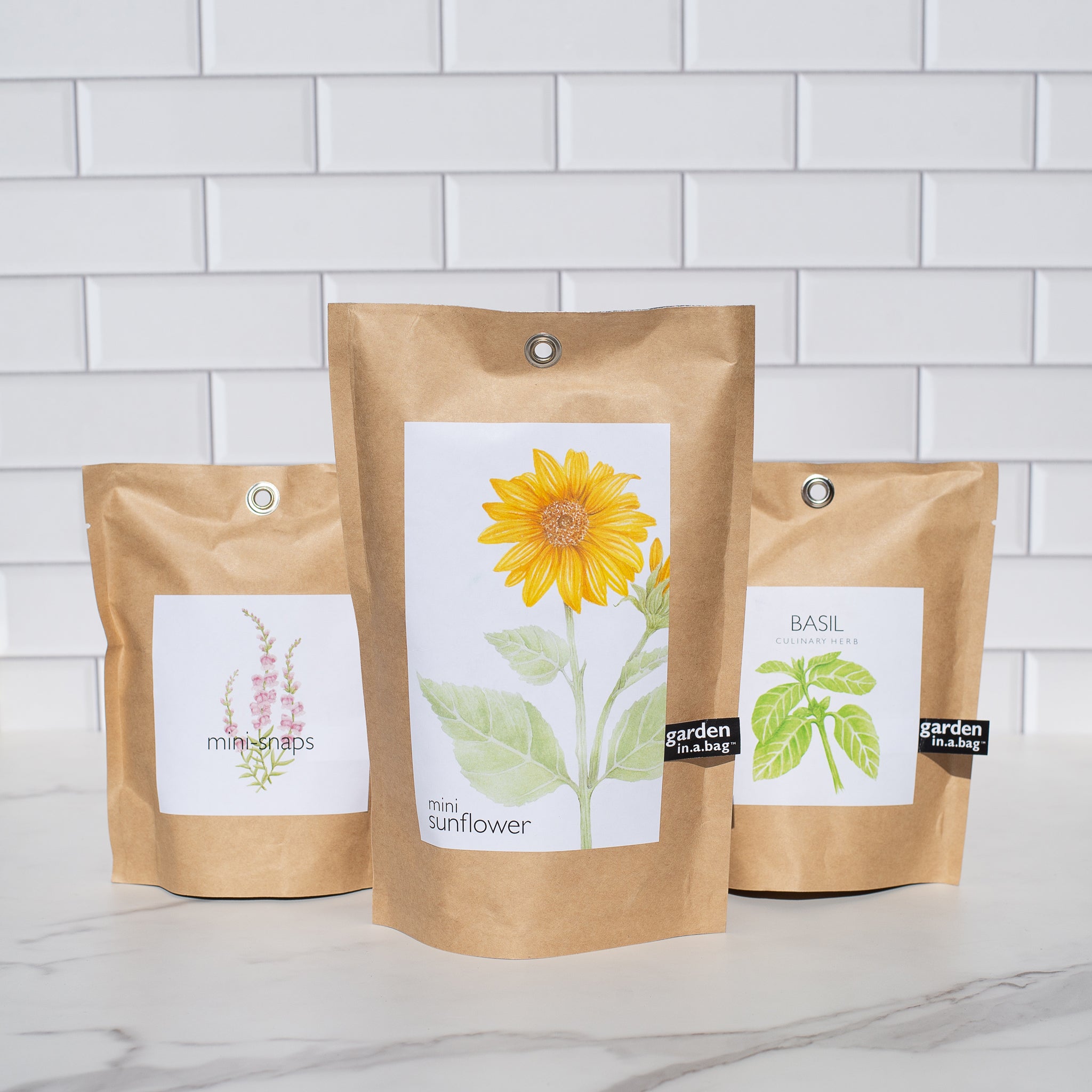 Three brown paper "garden in a bag" pouches are shown side-by-side. The left pouch is labeled "mini-snaps" the center is "mini sunflower" and the right is "BASIL".