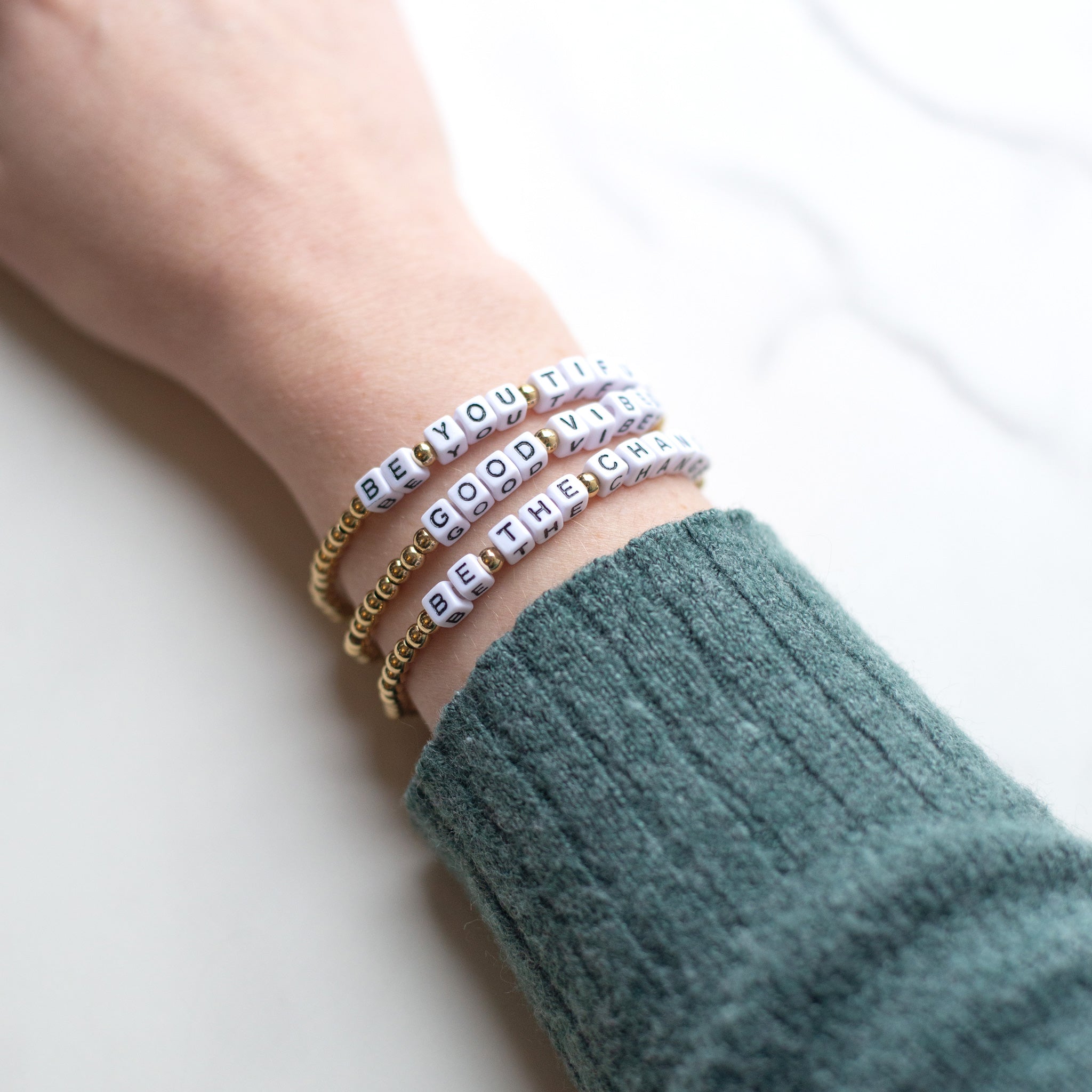 A hand wearing three 14K gold-plated word bracelets reading ‘Be You Tiful,’ ‘Be the Change,’ and ‘Good Vibes.’ Handmade in Bali from recycled stainless steel and plastic beads.
