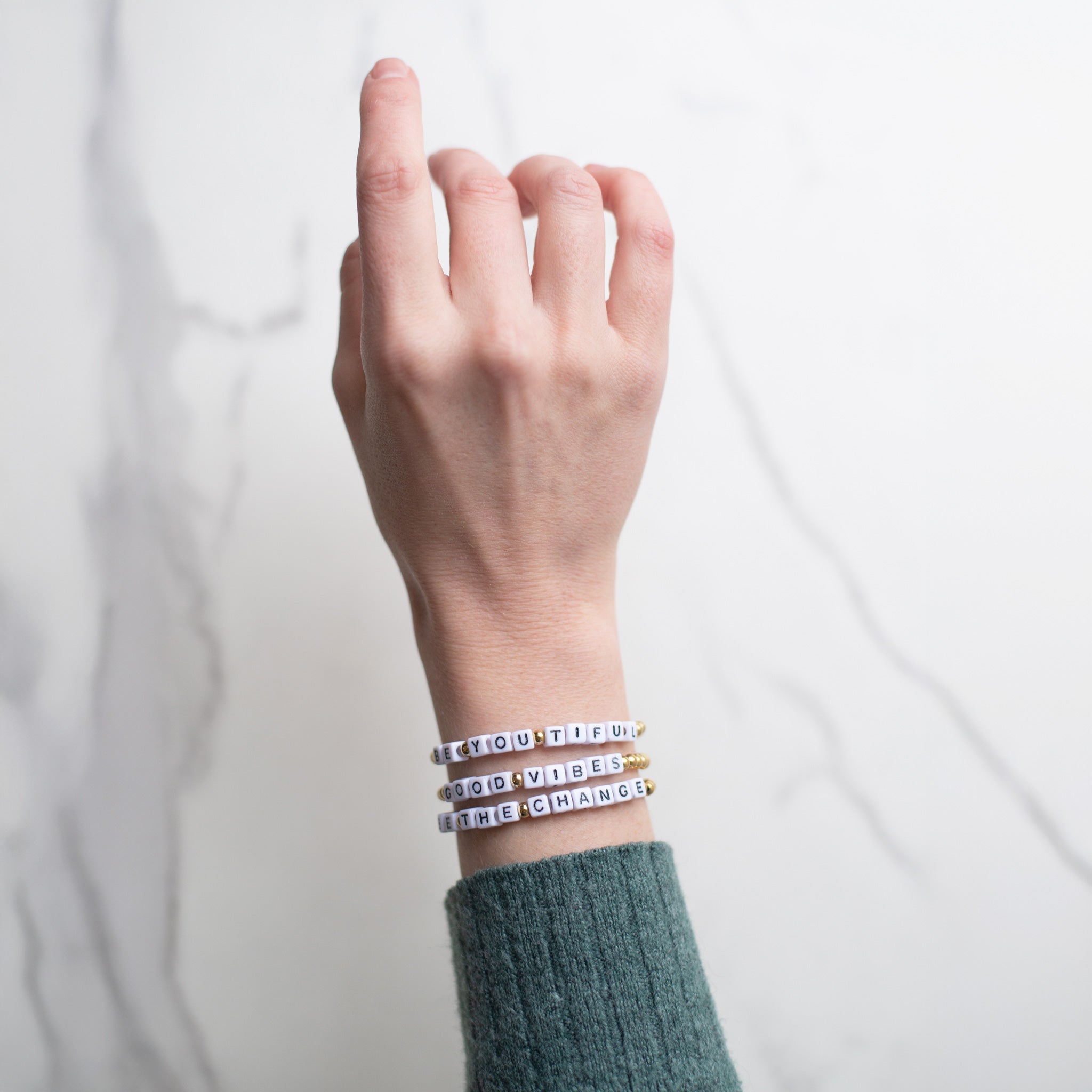 Close view of a hand adorned with four 14K gold-plated bracelets reading ‘Be the Change,’ ‘Good Vibes,’ and ‘Be You Tiful.’ Each handmade in Bali from recycled stainless steel and plastic beads