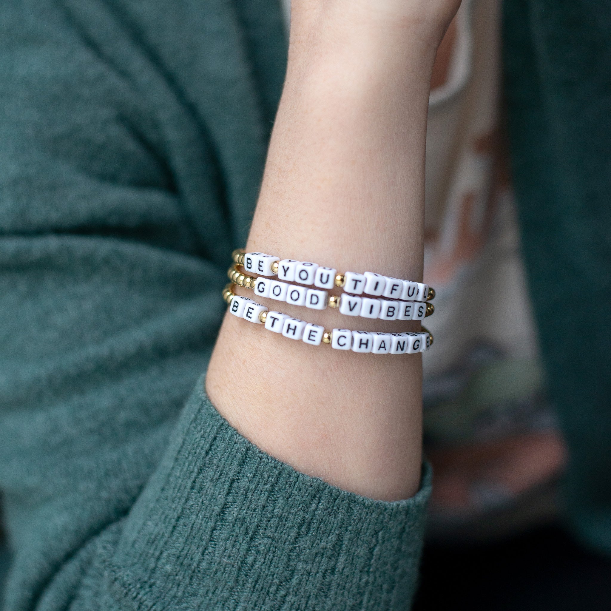 A hand wearing three 14K gold-plated word bracelets reading ‘Be You Tiful,’ ‘Be the Change,’ and ‘Good Vibes.’ Handmade in Bali from recycled stainless steel and plastic beads.