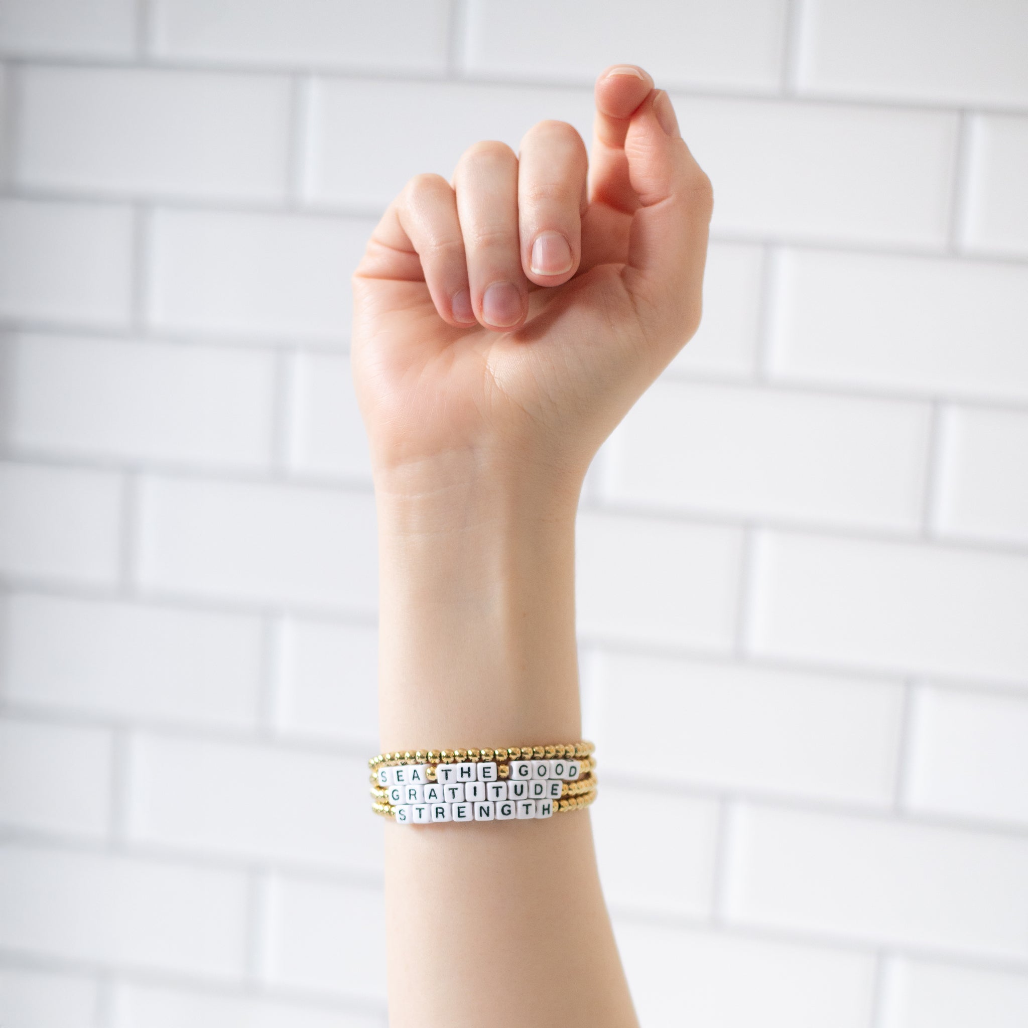 Hand wearing three 14K gold-plated word bracelets with the messages ‘Sea The Good,’ ‘Gratitude,’ and ‘Strength.’ Handmade in Bali using recycled materials, these water-resistant bracelets support ocean cleanup efforts.