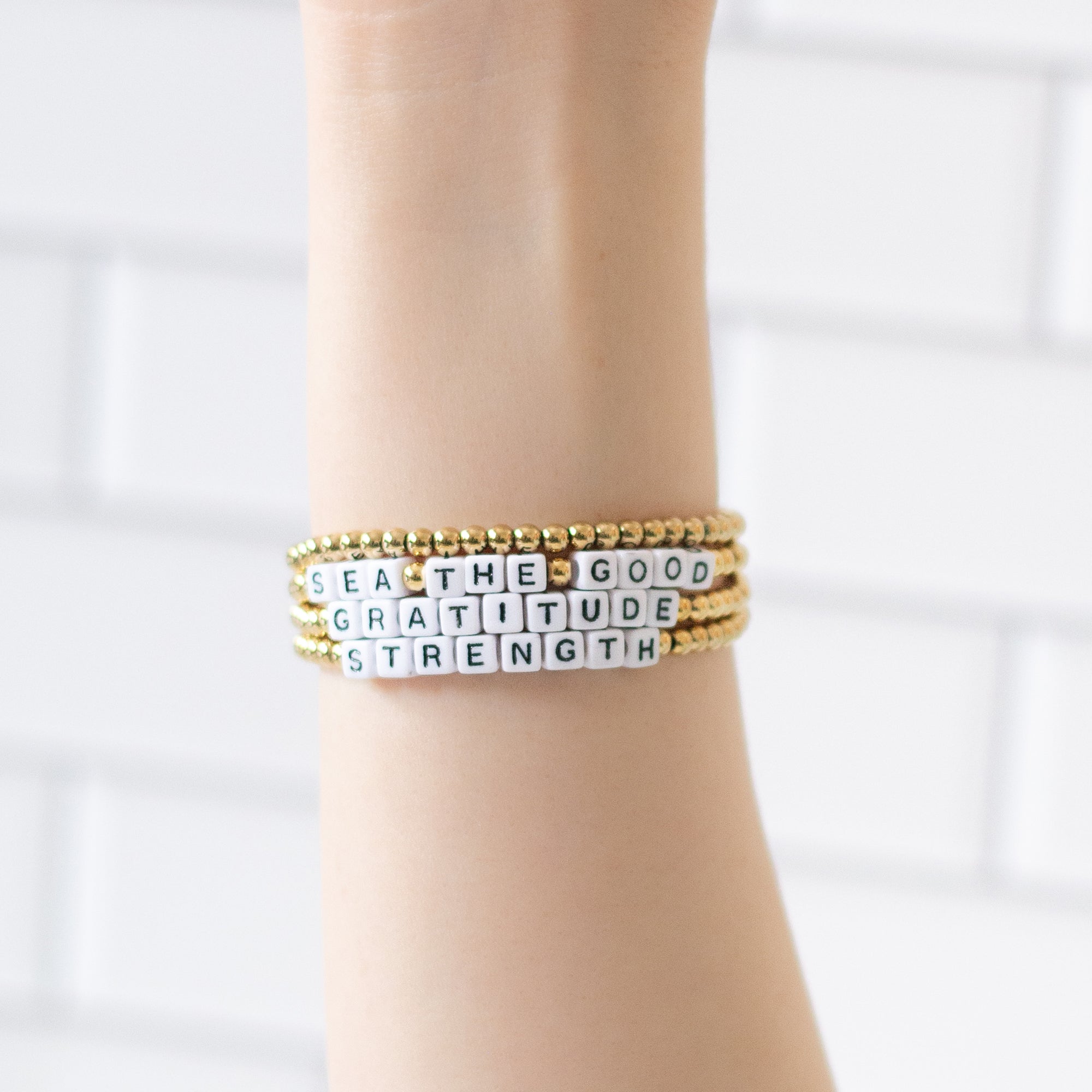 Hand wearing four 14K gold-plated word bracelets with the messages ‘Sea The Good,’ ‘Gratitude, and ‘Strenght,’ . Handmade in Bali using recycled materials, these water-resistant bracelets support ocean cleanup efforts.