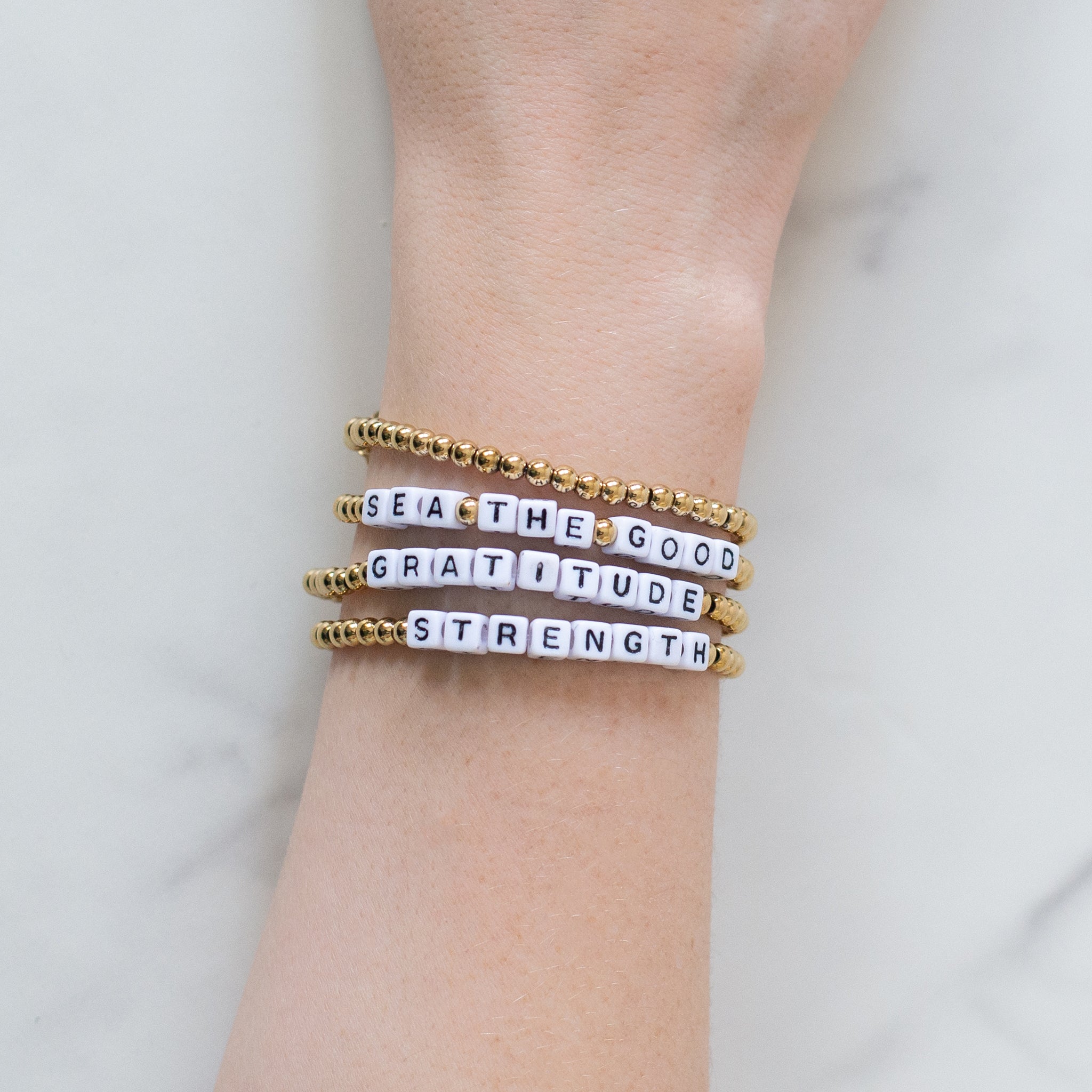 Hand wearing three 14K gold-plated word bracelets reading ‘Sea the Good,’ ‘Strength,’ and ‘Gratitude.’ Handmade in Bali from recycled materials, water-resistant, and supporting ocean cleanup efforts.