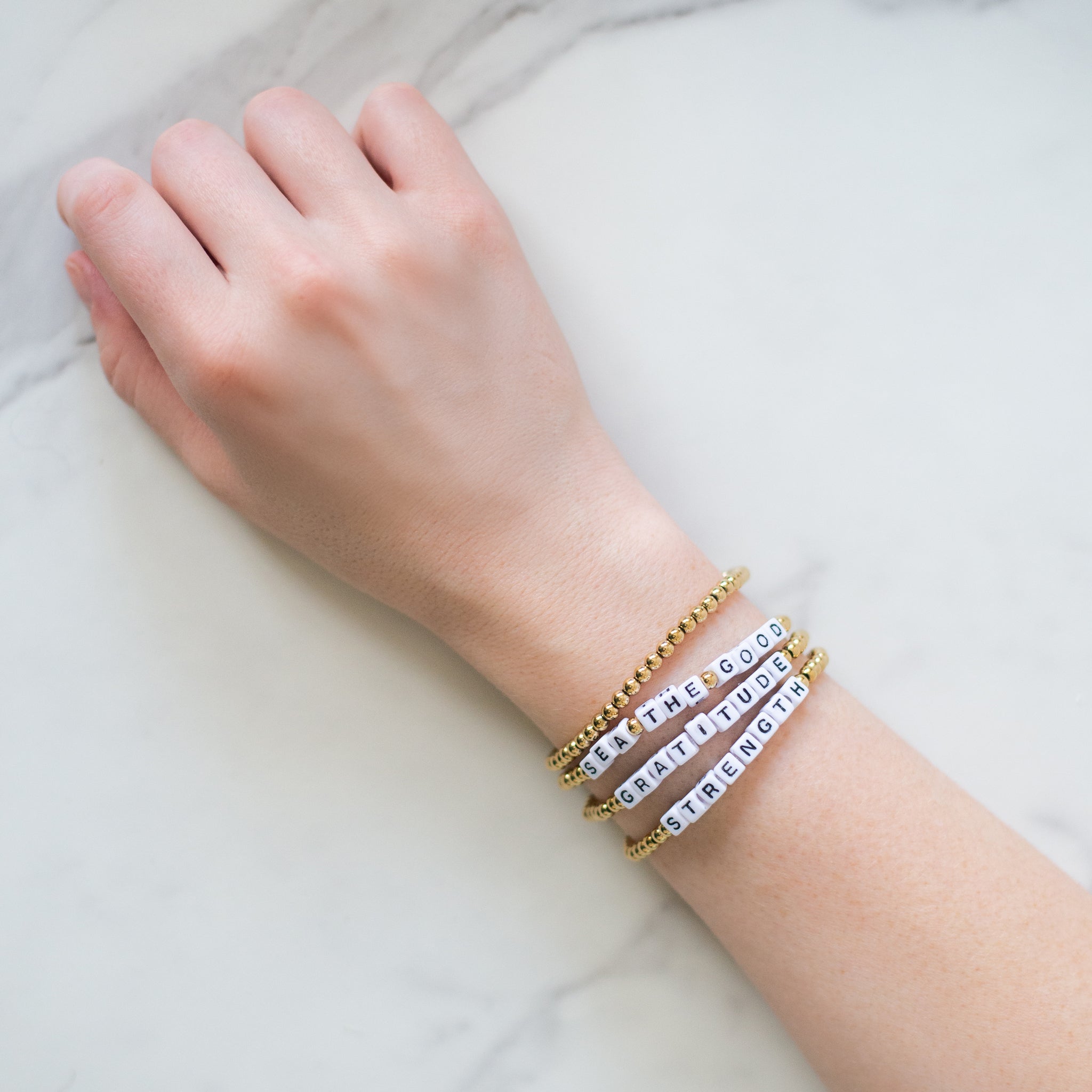 Hand wearing three 14K gold-plated word bracelets reading ‘Sea the Good,’ ‘Strength,’ and ‘Gratitude.’ Handmade in Bali from recycled materials, water-resistant, and supporting ocean cleanup efforts.