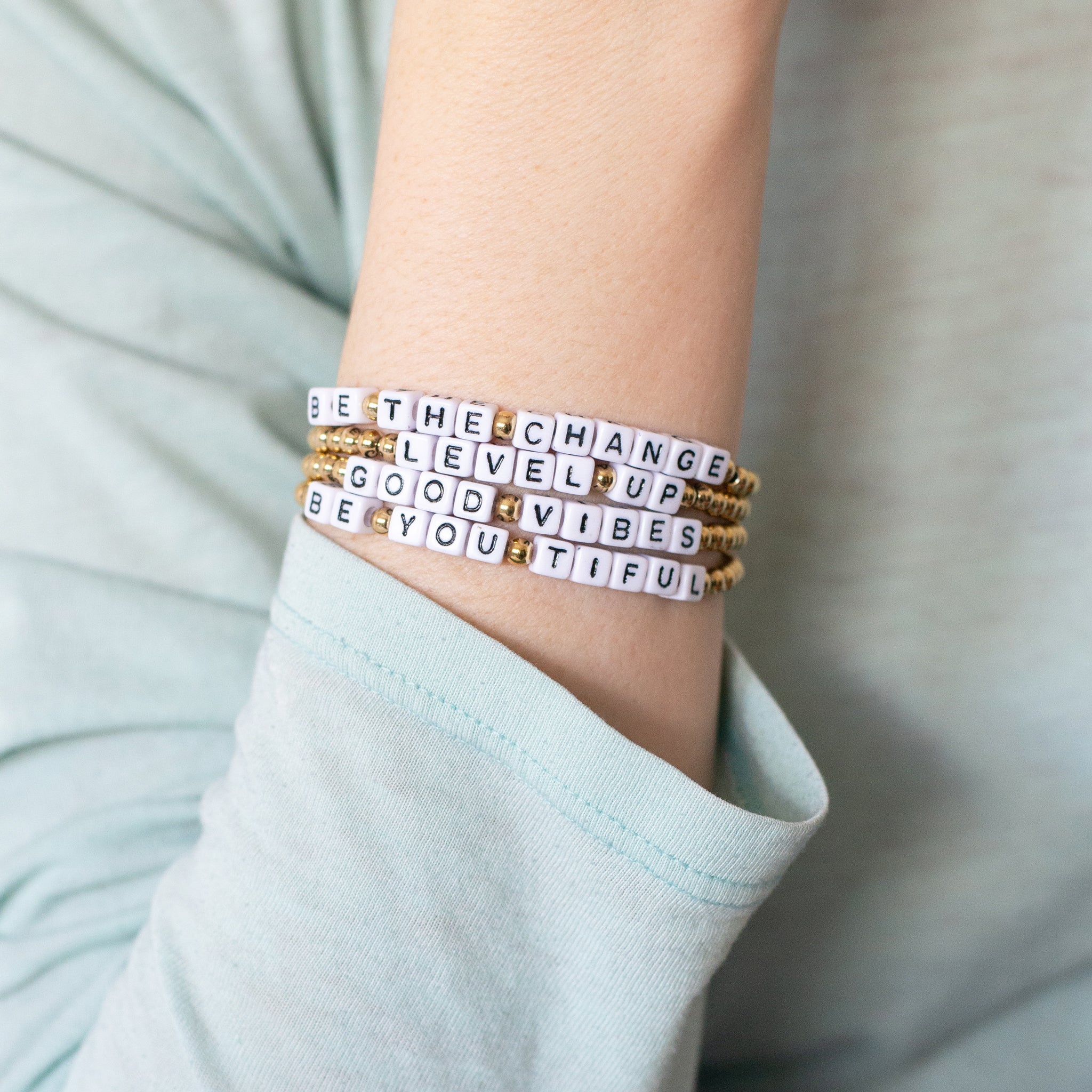 Hand wearing four 14K gold-plated word bracelets with the messages ‘Level Up,’ ‘Be the Change,’ ‘Good Vibes,’ and ‘Be You Tiful.’ Handmade in Bali from recycled materials, water-resistant, and supporting ocean cleanup.