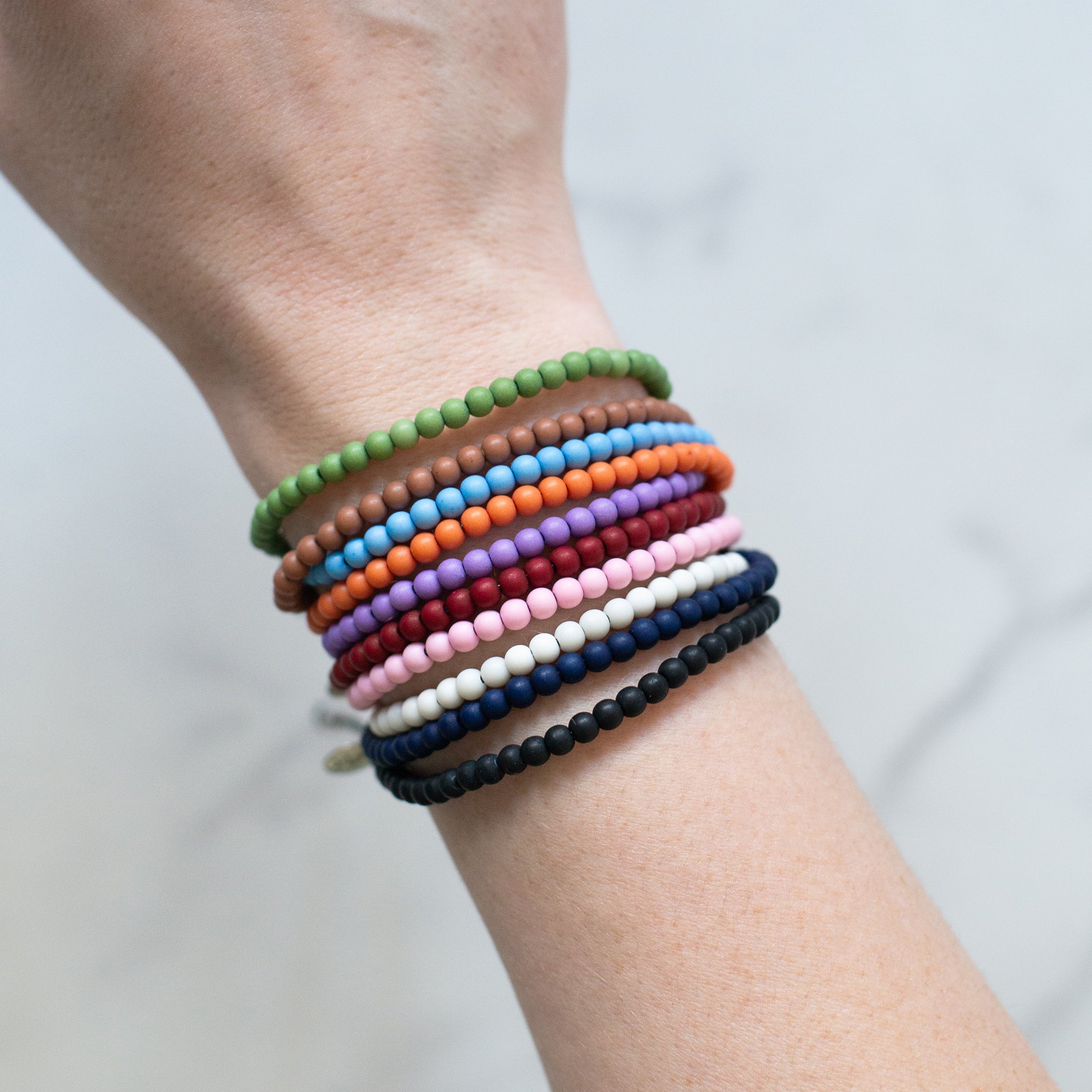 Stack of recycled ocean plastic bead bracelets in various colors, worn on a wrist. Adjustable cotton cords and stainless steel charms, handmade in Bali and water-safe.