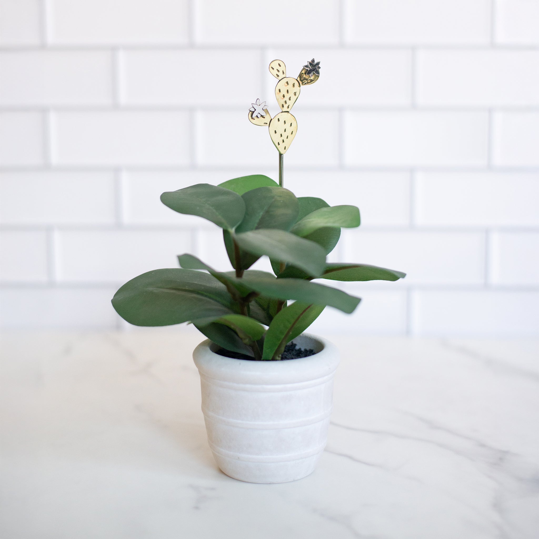 Cactus-shaped plant stake made from alpaca silver and brass, displayed in a small potted plant against a white tiled background.