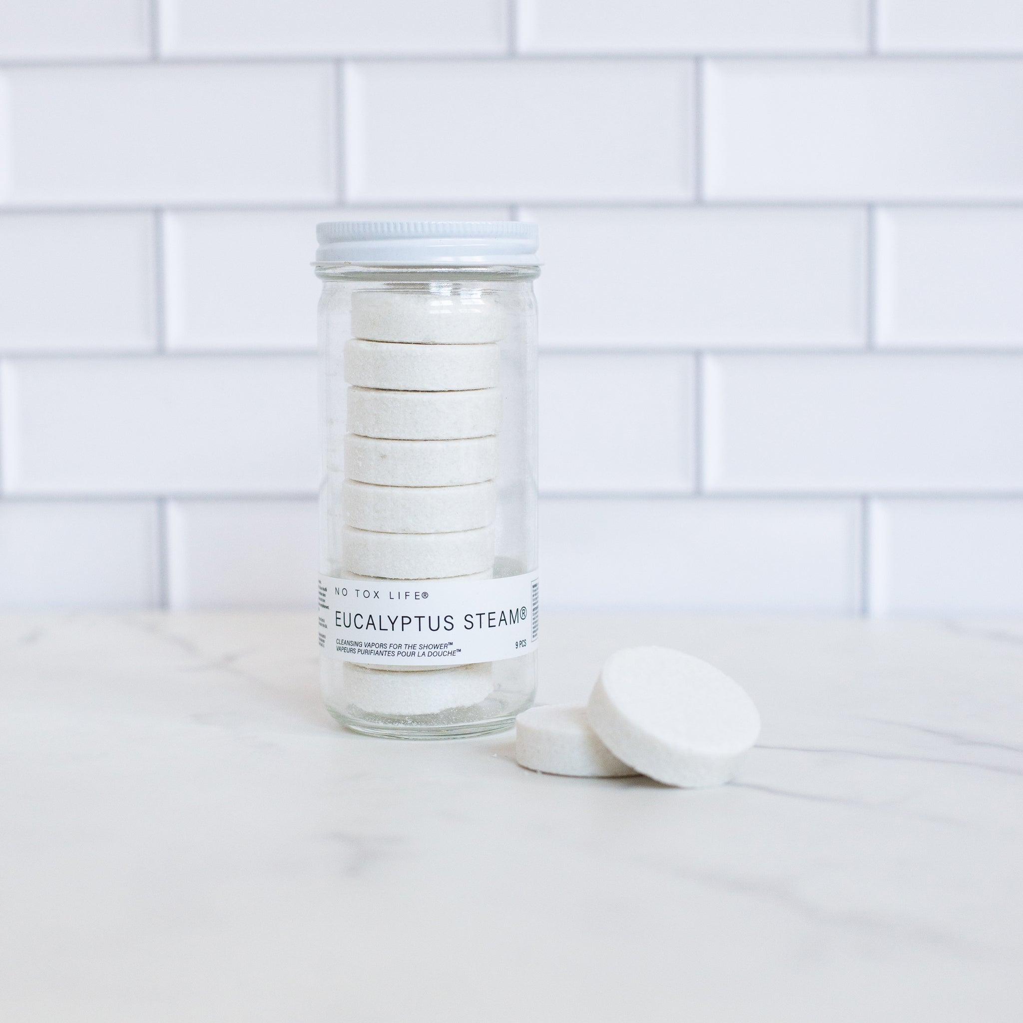 A tall glass jar filled with eight white eucalyptus steam discs, and two more discs are stacked next to it. The jar has a white lid and a "No Tox Life" brand label.