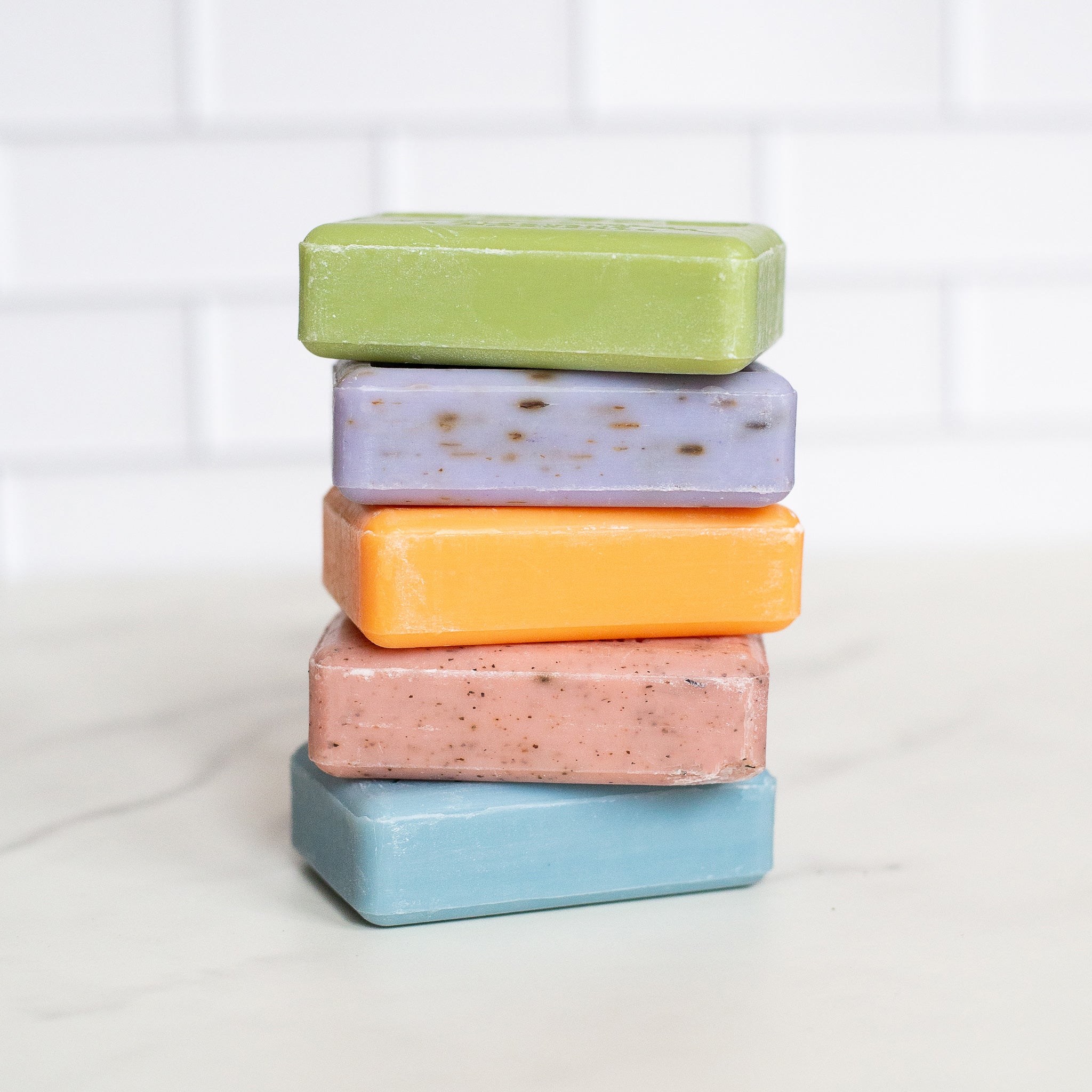 A stack of five rectangular french soap bars rests on a white counter. From top to bottom the bars are green, purple, orange, pink with speckles and pale blue.