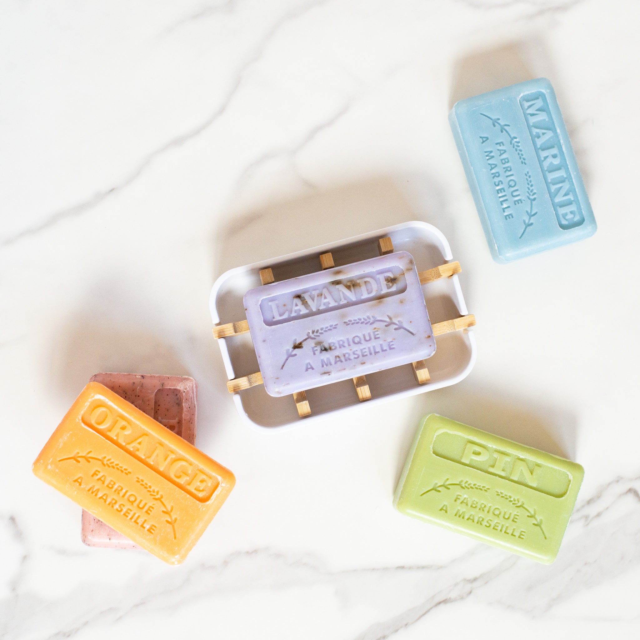 Five French soap bars sit on a white marble surface, including one lavender soap on a white dish with bamboo slats. The other soaps are orange, pink, light blue and green.