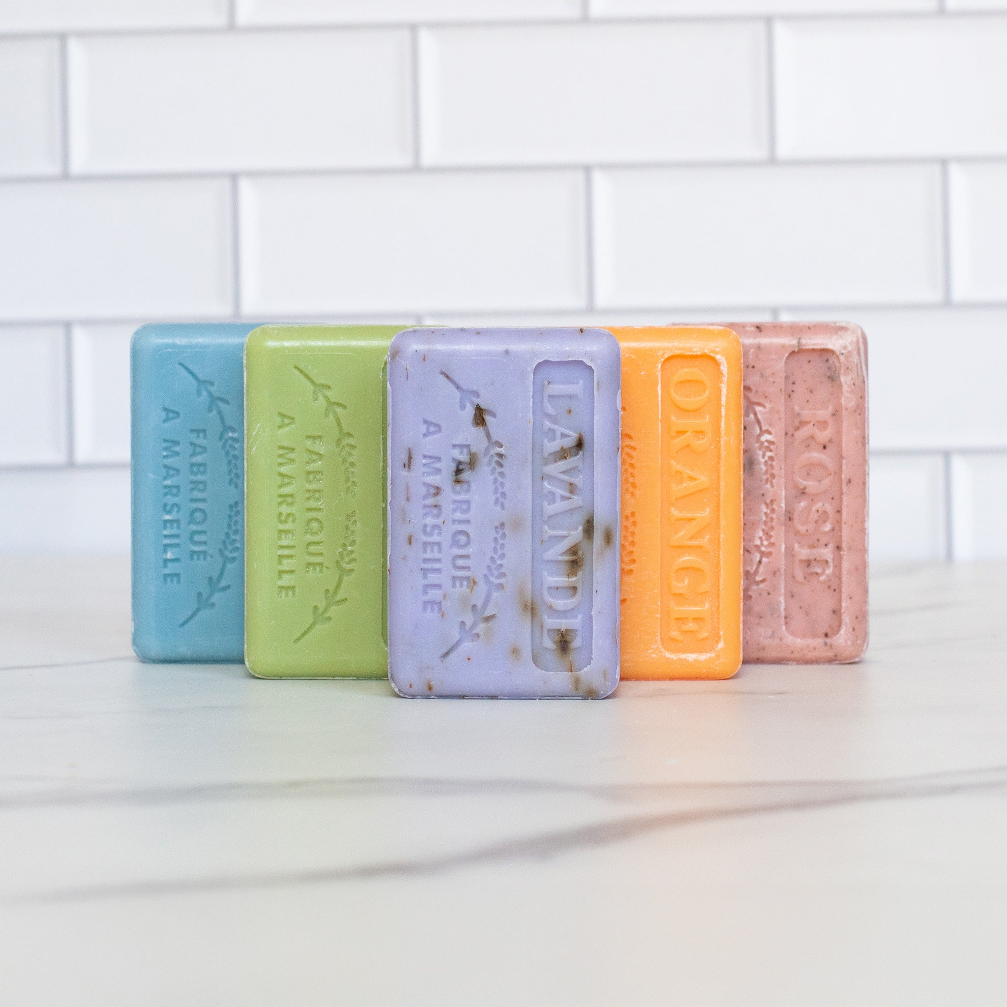 Five French soap bars stand in a staggered row on a white marble counter. The soaps are lavender orange pink green and blue each with their name embossed on the surface.