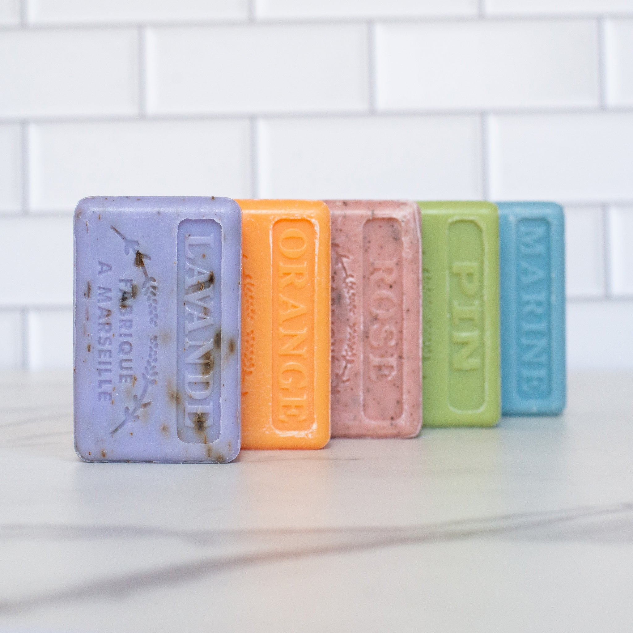 A lineup of five French soap bars stands upright on a white marble counter. The colors, from left to right, are lavender orange pink green and blue, each with their name embossed on the surface.