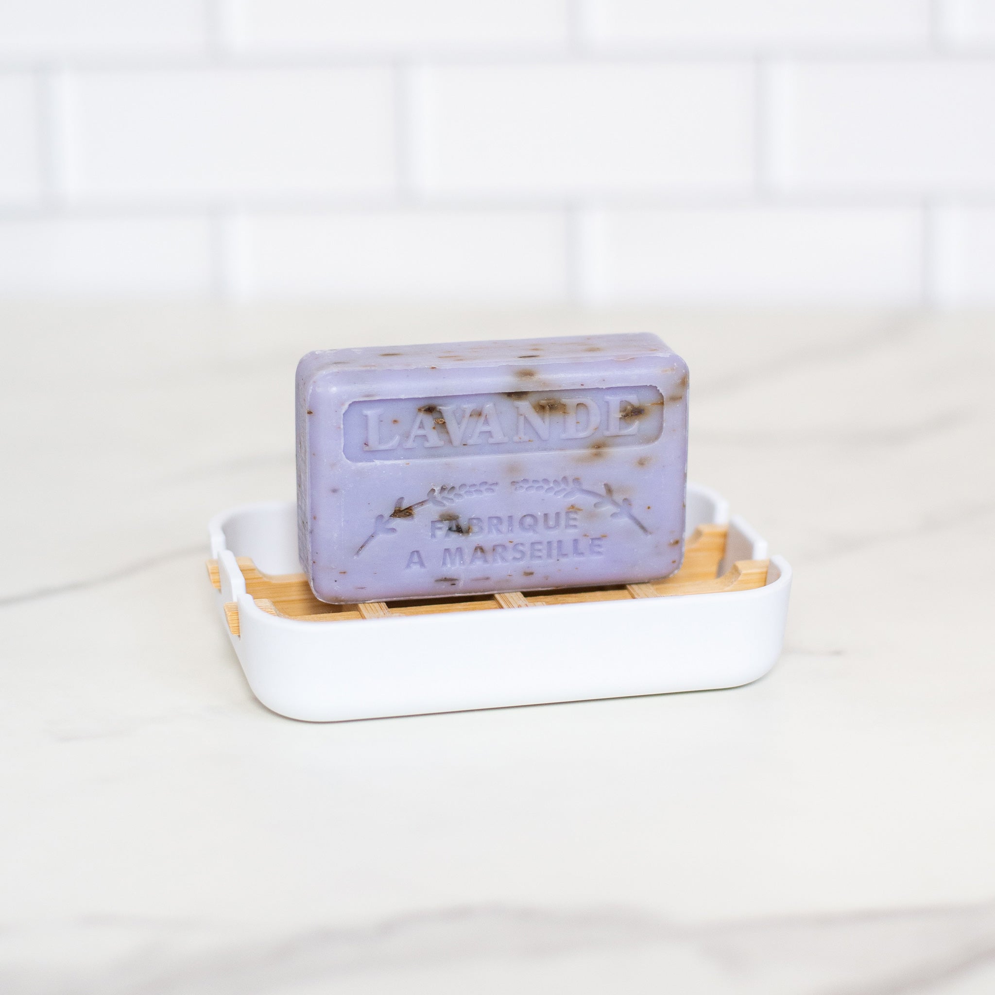 A "Lavande" French soap bar is on a white dish with bamboo slats on a white marble surface. It has embossed text and dark speckles.