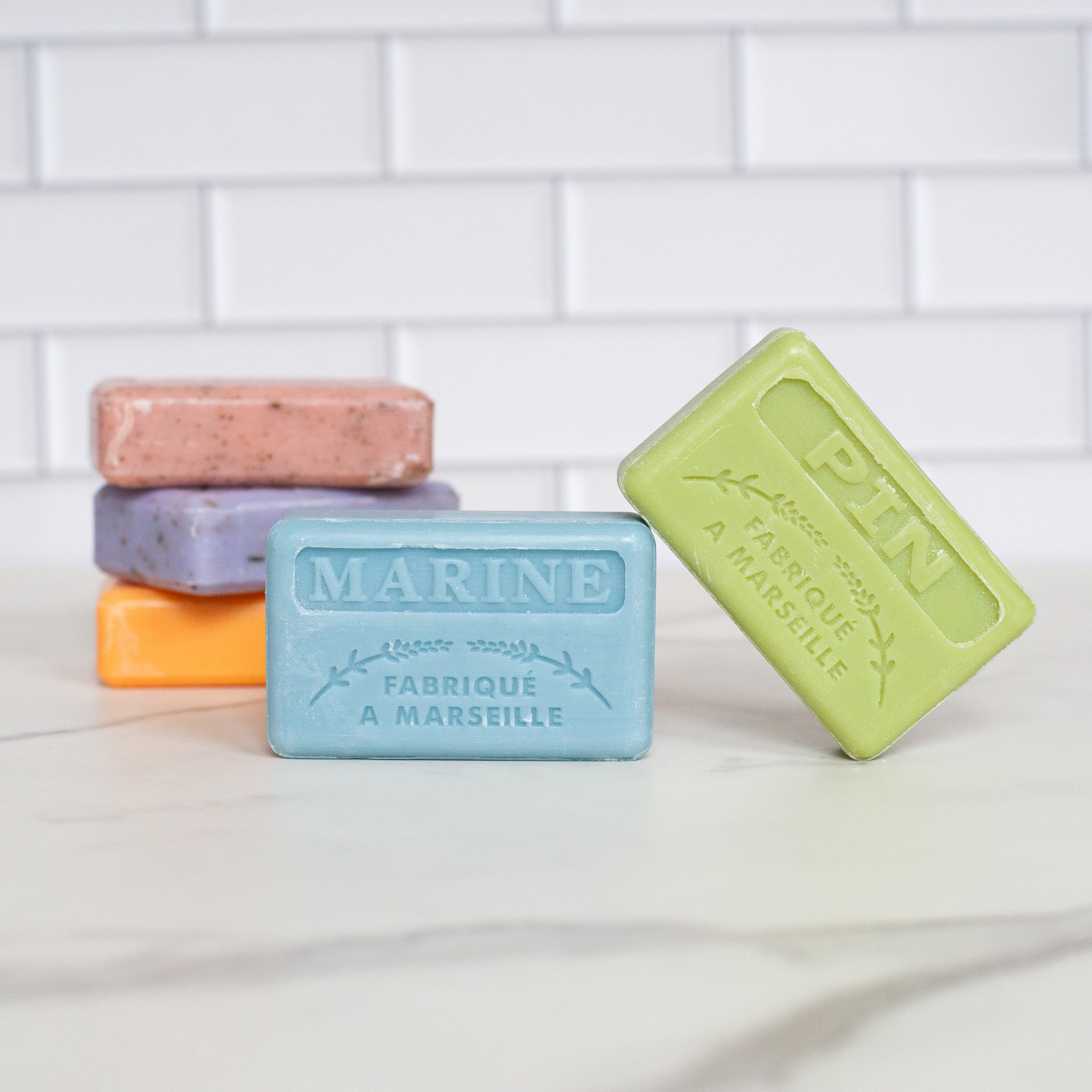 A group of five soap bars is on a white counter. A blue soap bar with "Marine" embossed on it and a green "Pin" bar are propped in front of a stack of pink, purple and orange soaps.