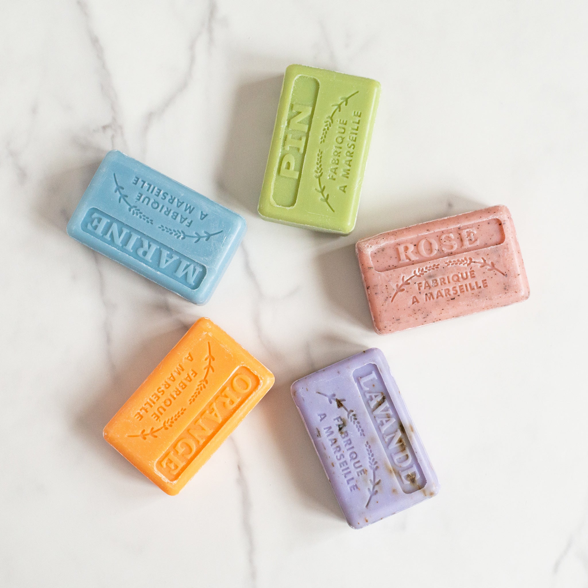 Five French soap bars are arranged in a circle on a white marble counter. The colors are green (Pin), blue (Marine), pink (Rose), orange (Orange) and lavender (Lavande).