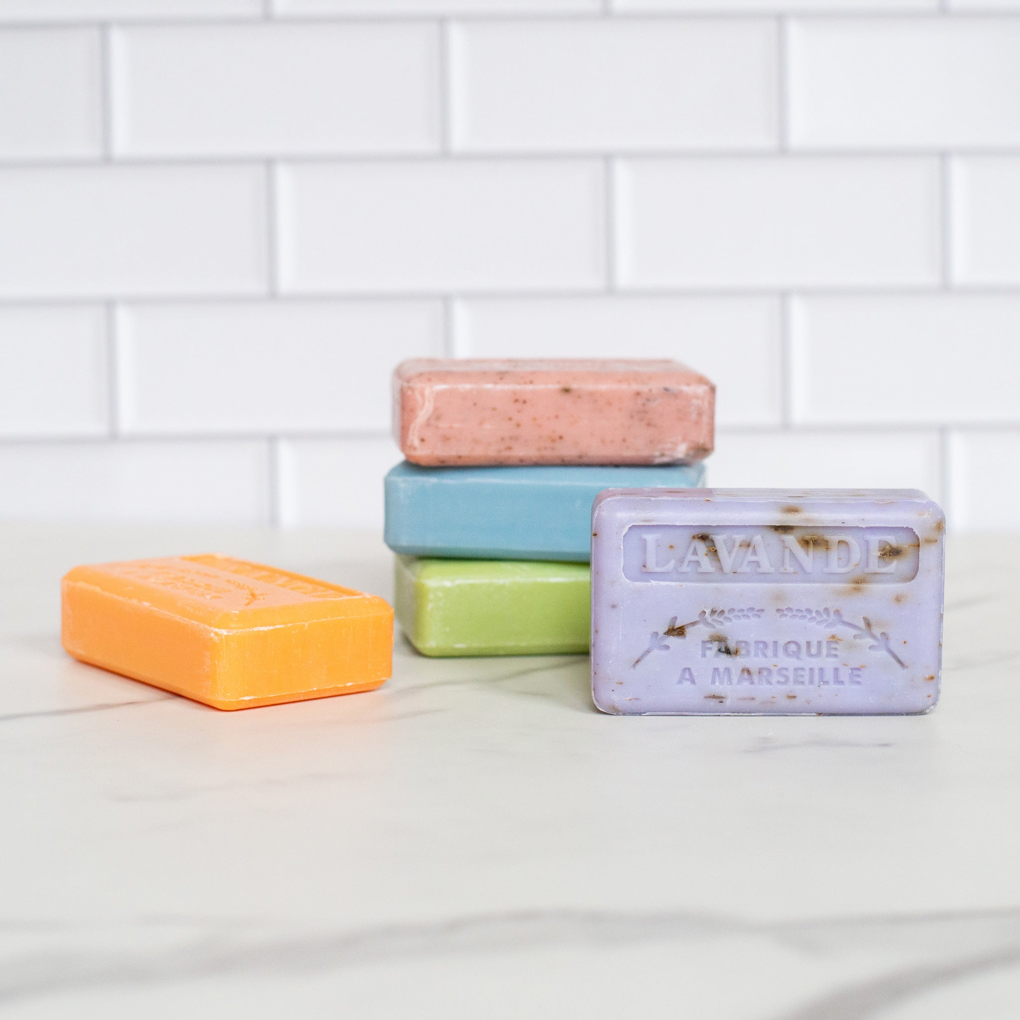 An arrangement of French soap bars is on a white counter. A purple soap is in the foreground with an orange soap beside it. Behind them is a stack of pink, blue and green bars.