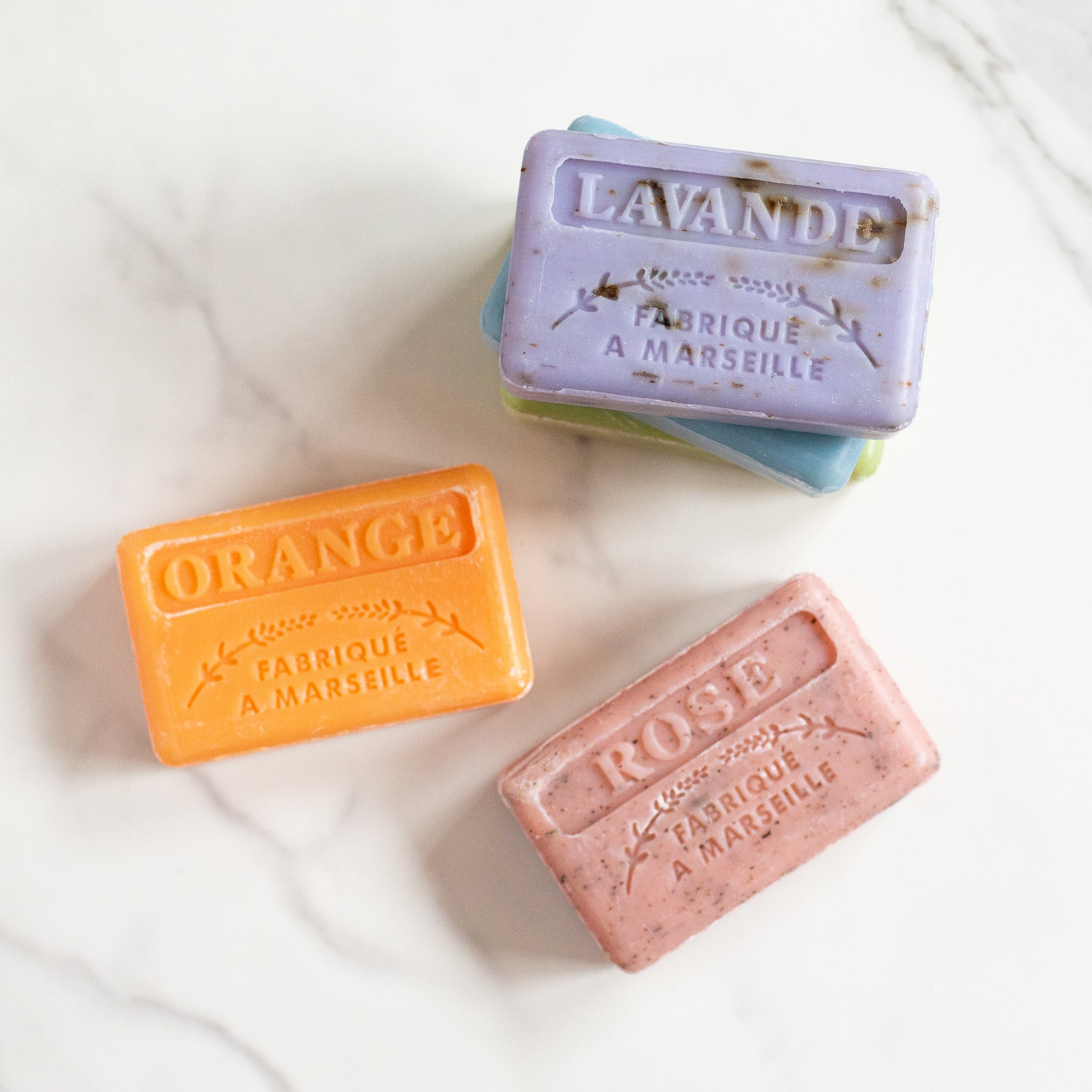 Four French soap bars are on a white marble counter. An orange "Orange" soap and a pink "Rose" soap are beside a stack of a blue bar and a lavender "Lavande" bar.