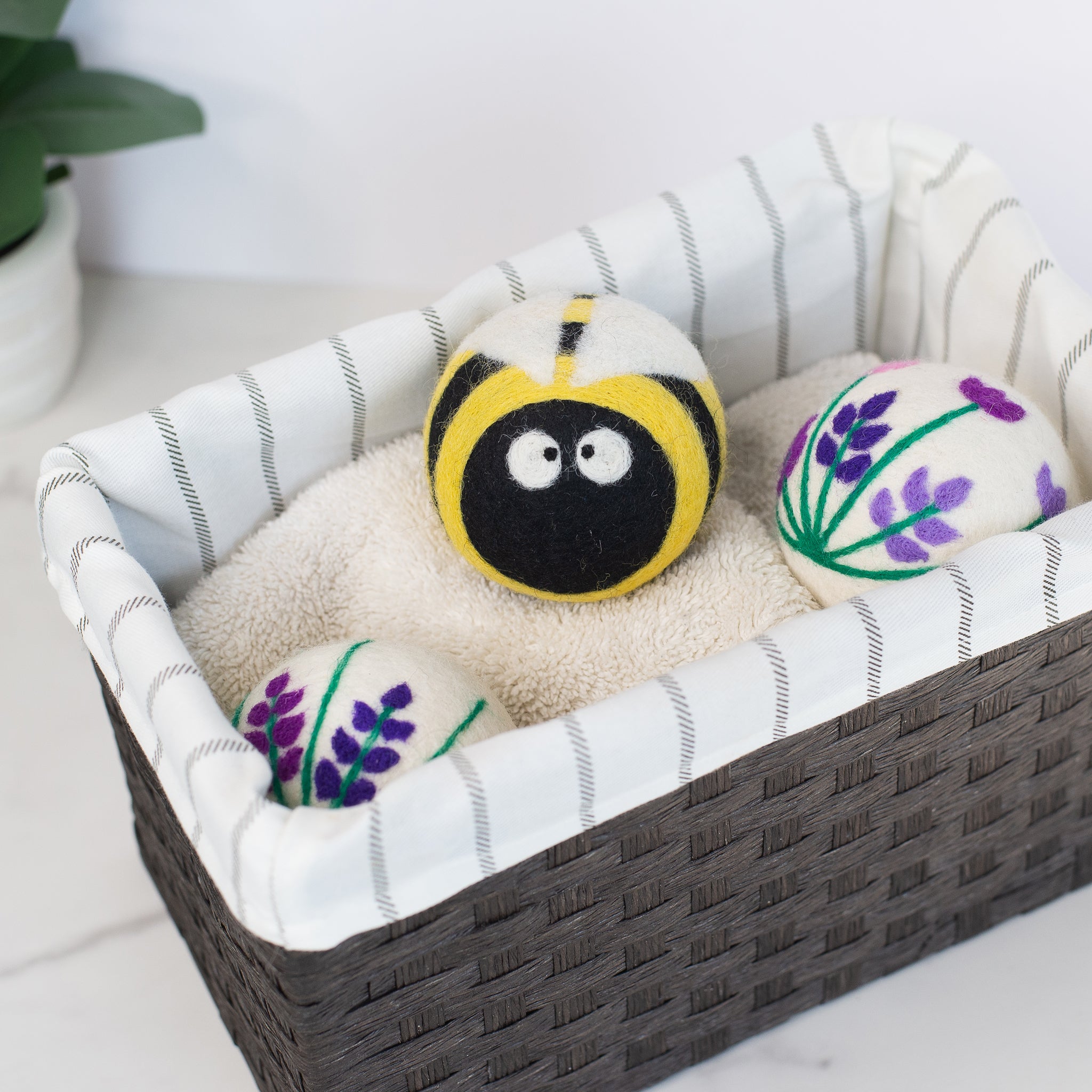 A set of three adorable Friendsheep sustainable goods wool dryer balls decorated with a cute bee and two purple floral designs are nestled in a wicker basket on a striped cloth liner.