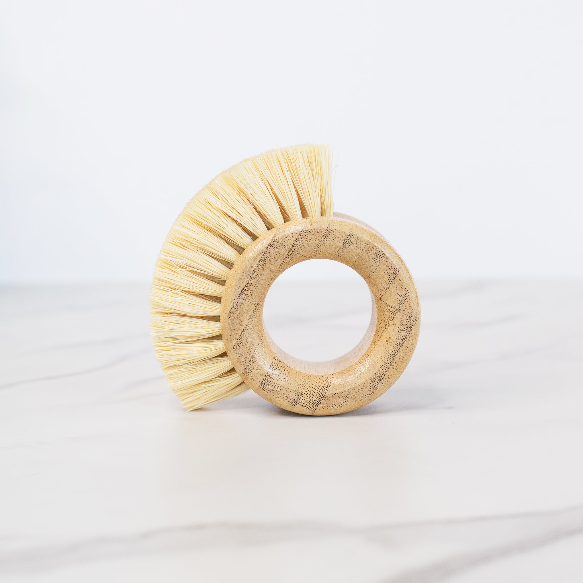 A light-colored bamboo vegetable brush is shaped like a thick ring. It has light blonde bristles that fan out in a semi-circle from the upper half of the ring.