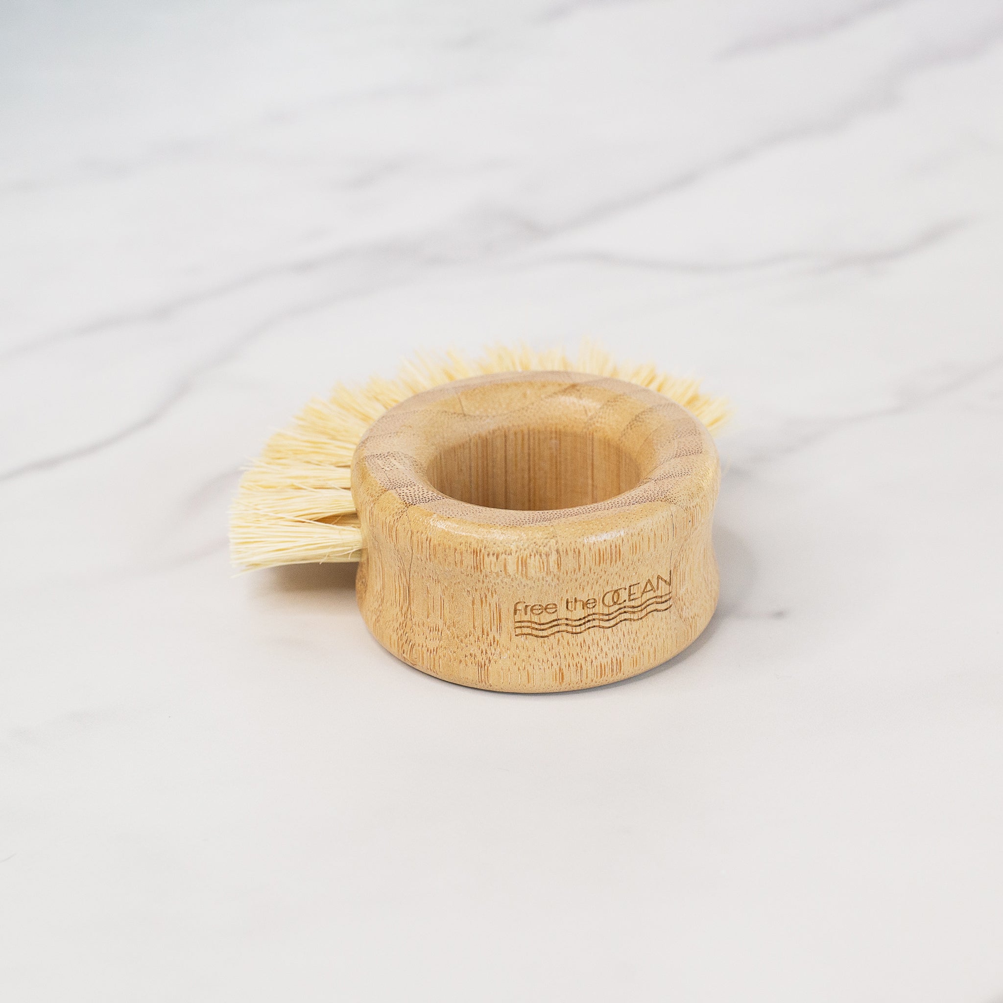 A round scrub brush with a thick bamboo ring handle. It has light-colored bristles and the words "free the OCEAN" with a wave logo are engraved on its side.