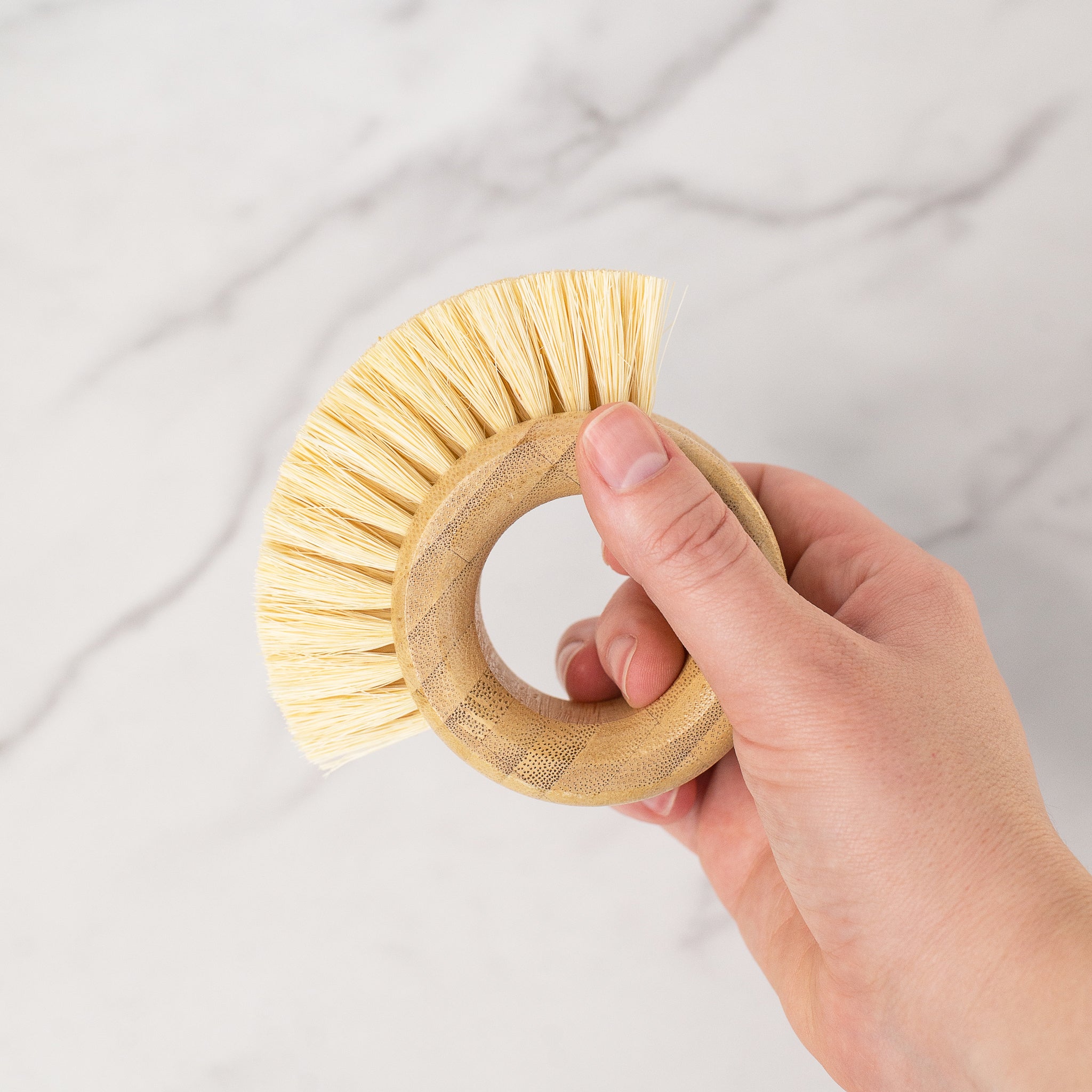 A hand holds a ring-shaped bamboo veggie brush against a white marble background. The brush has light blonde bristles on its outer curve.