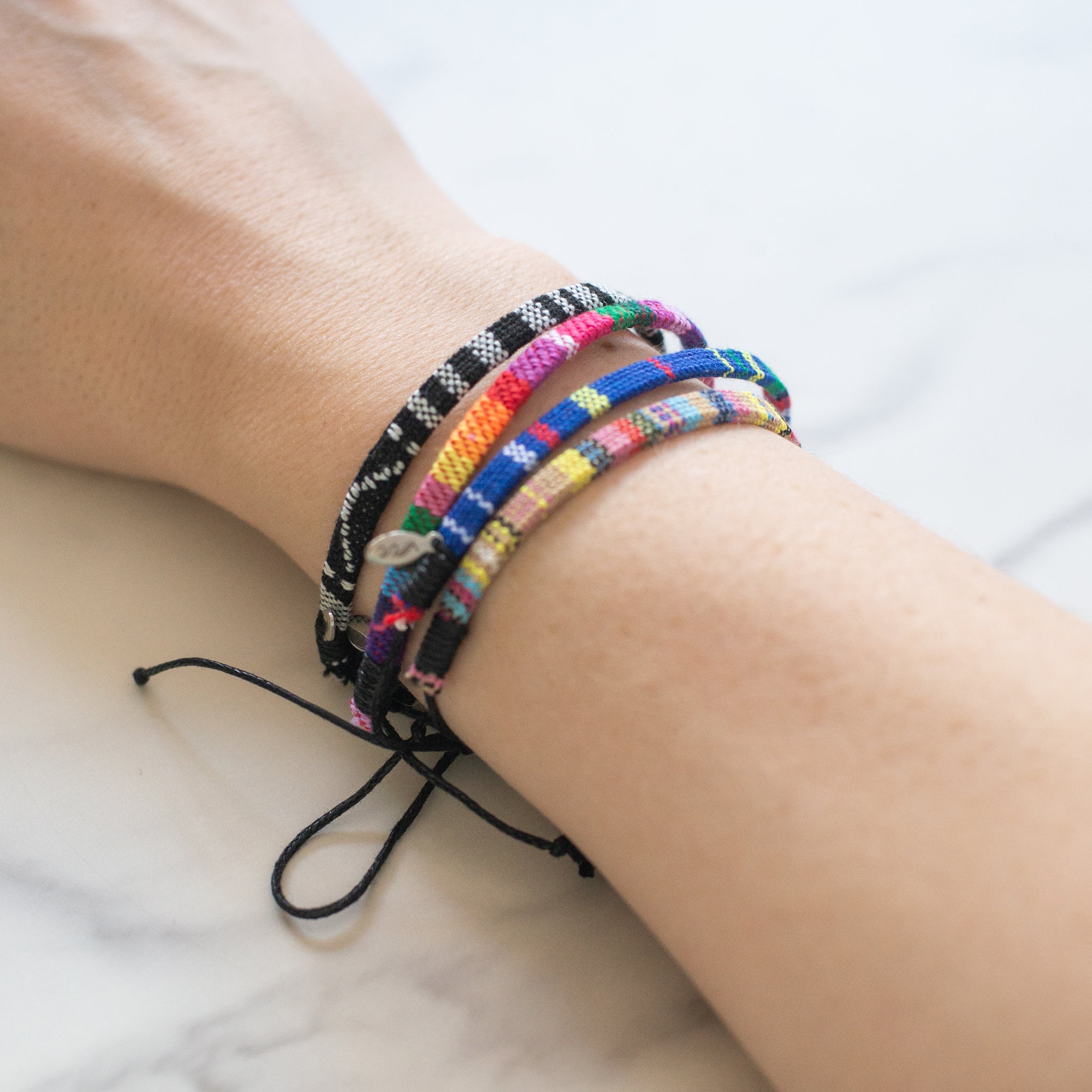 Stack of woven bracelets in various colorways, worn on wrist. Adjustable and water-resistant with recycled stainless steel charms. Handmade in Bali to support ocean cleanup efforts.