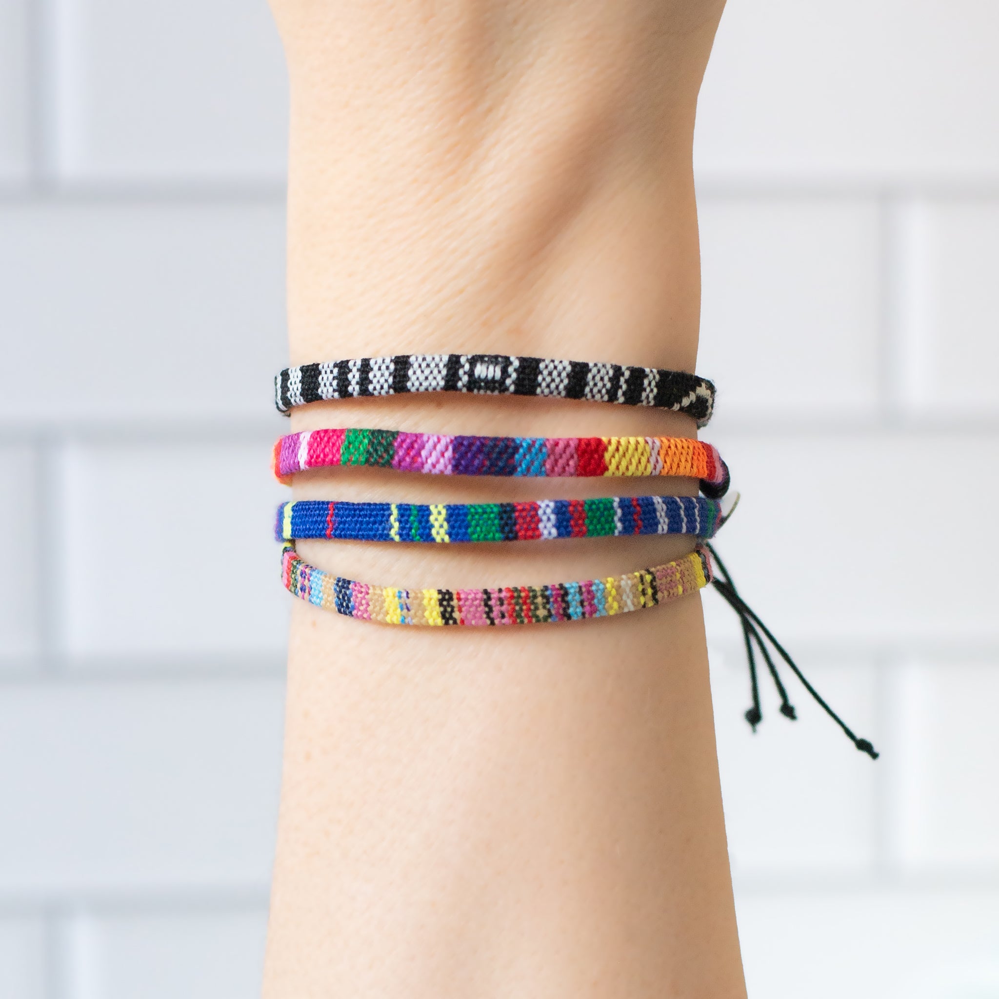 Stack of colorful woven bracelets on a wrist, featuring various patterns and shades. Handmade in Bali with adjustable cotton-blend cords and recycled stainless steel charms. Each bracelet helps remove one pound of trash from waterways.
