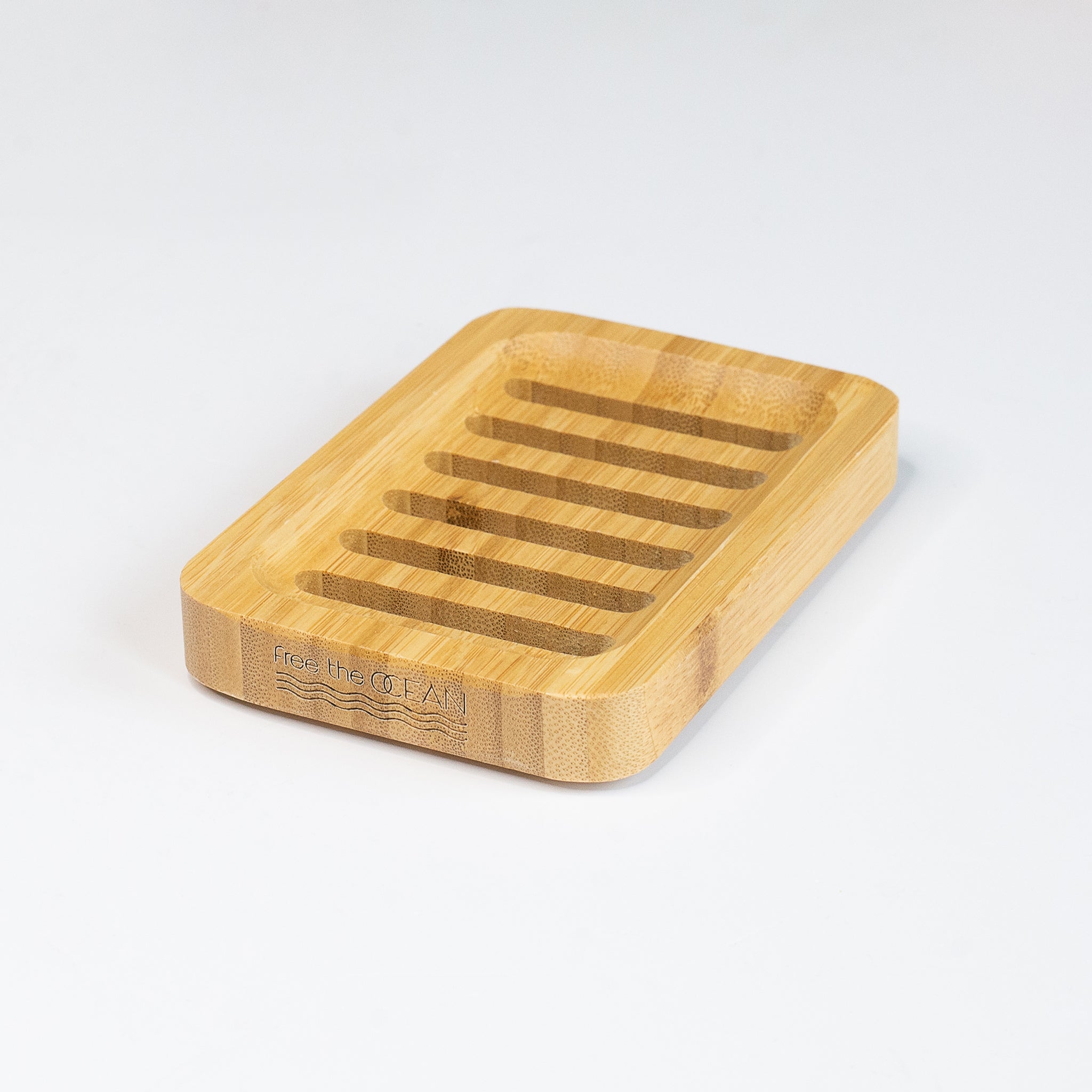 A rectangular bamboo soap dish is shown with a Free the OCEAN logo etched into its side. The dish has several parallel slats carved into its surface for drainage.