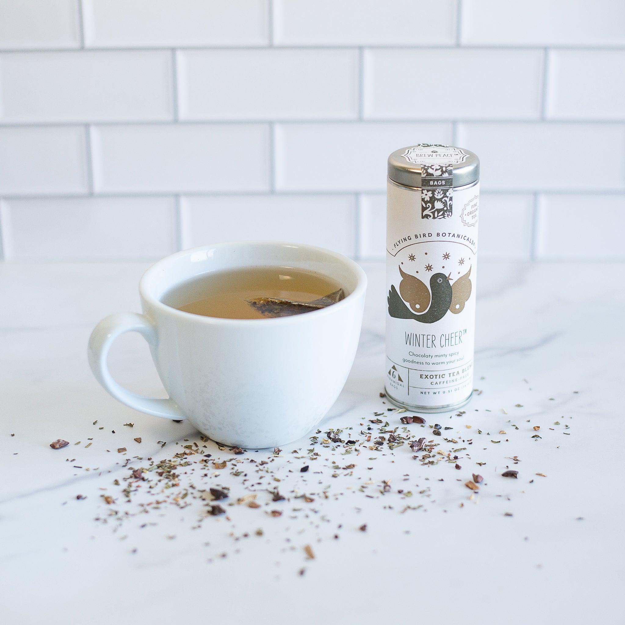 A cup of tea sits next to a tin of "Winter Cheer" tea from Flying Bird Botanicals. The label on the tin features two hummingbirds and the words "Chocolate minty goodness to warm your day". Loose tea leaves are scattered on the white marble counter.
