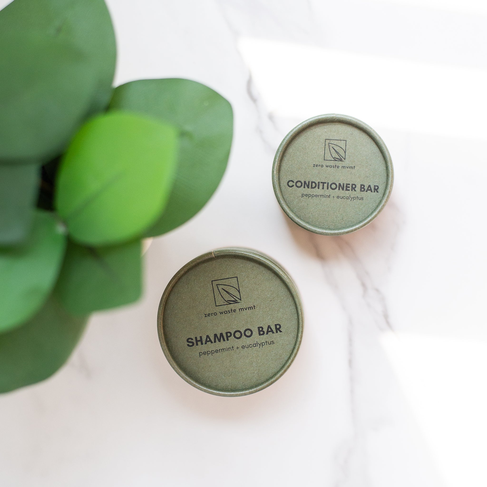 Two round green "zero waste mvmt" containers for shampoo and conditioner bars sit on a white marble surface next to a green plant. Both are peppermint + eucalyptus.
