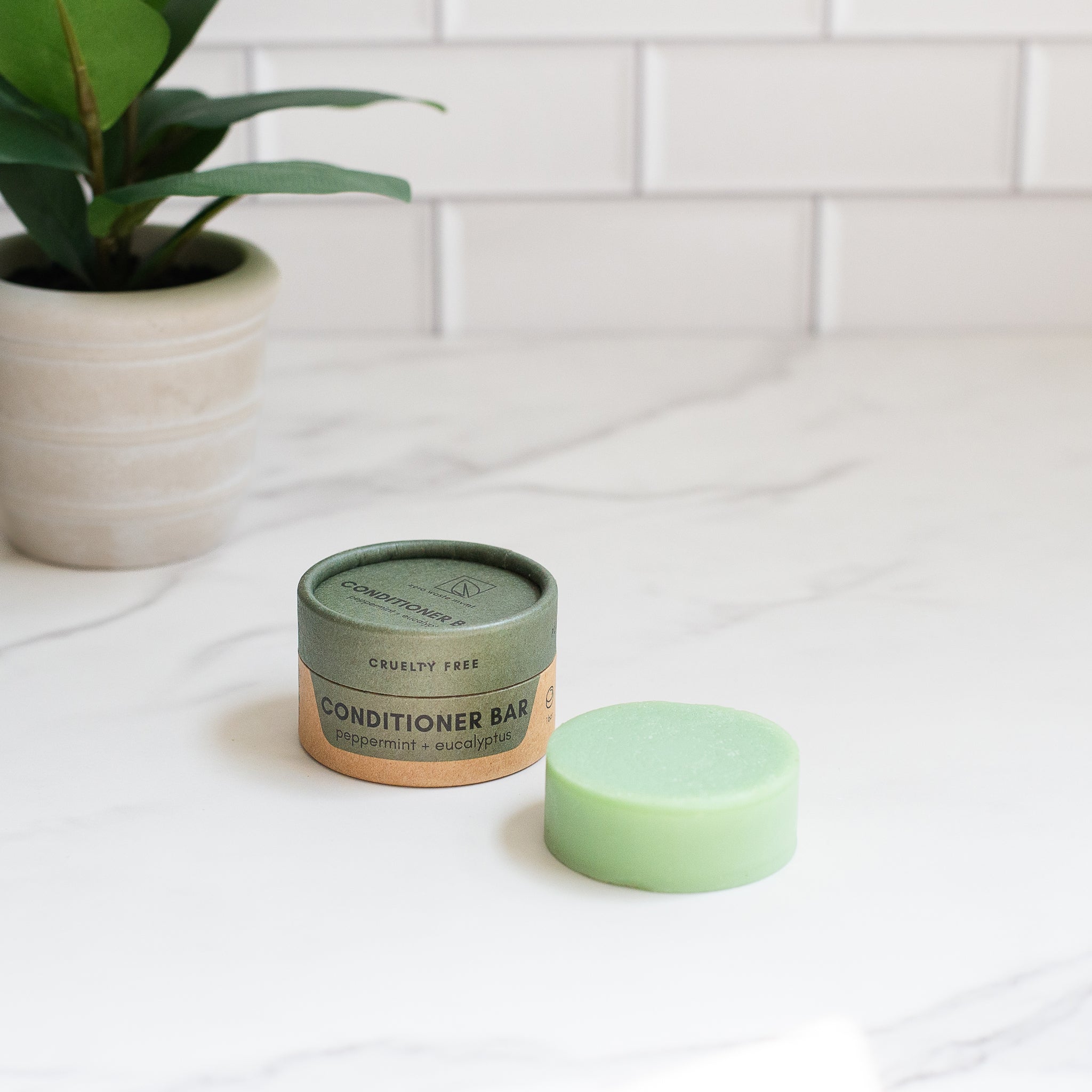 A round "CRUELTY FREE" conditioner bar rests next to its "peppermint + eucalyptus" container on a white marble surface.