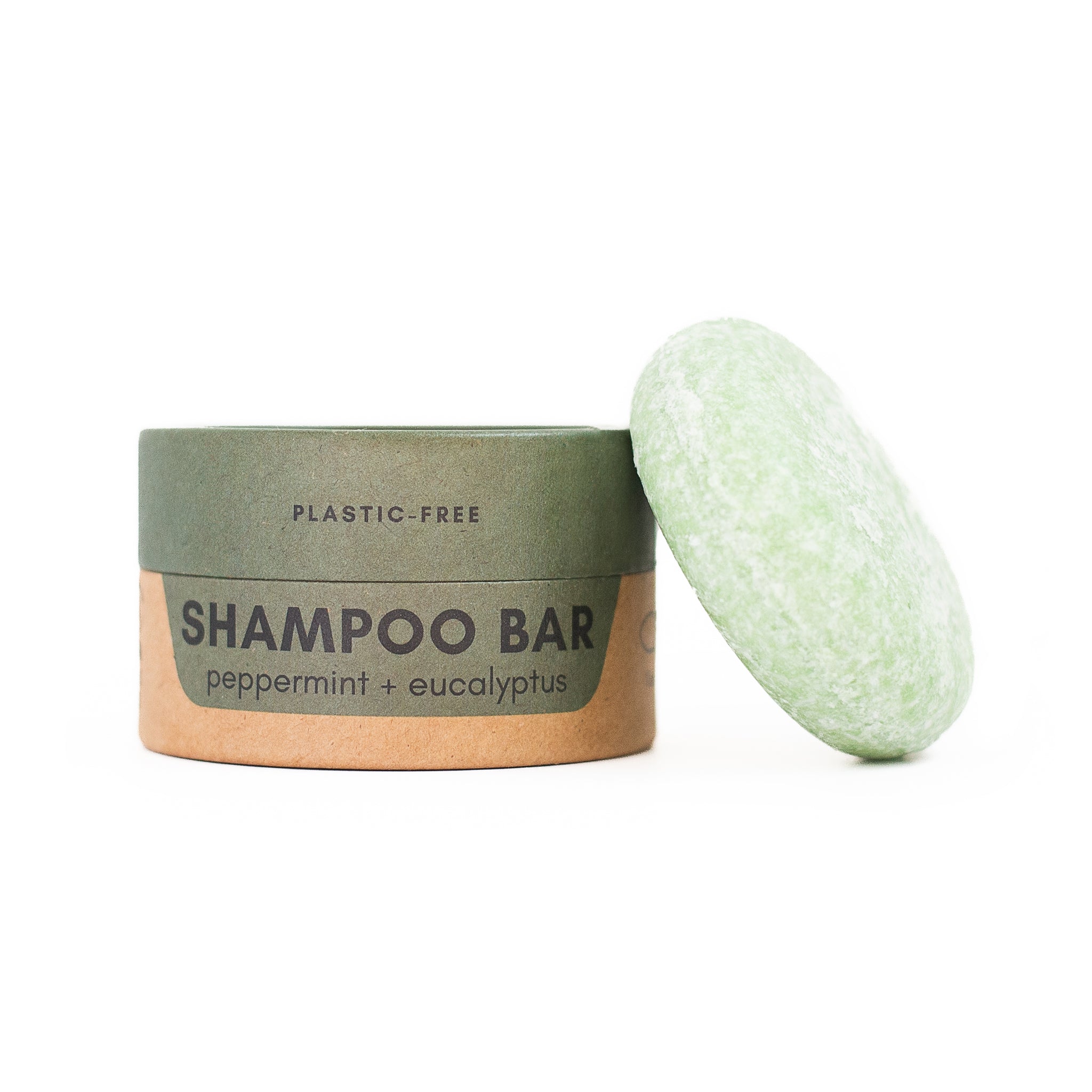 A two-toned cardboard container labeled "PLASTIC-FREE SHAMPOO BAR" with peppermint and eucalyptus stands next to a speckled pale green oval-shaped solid shampoo bar.