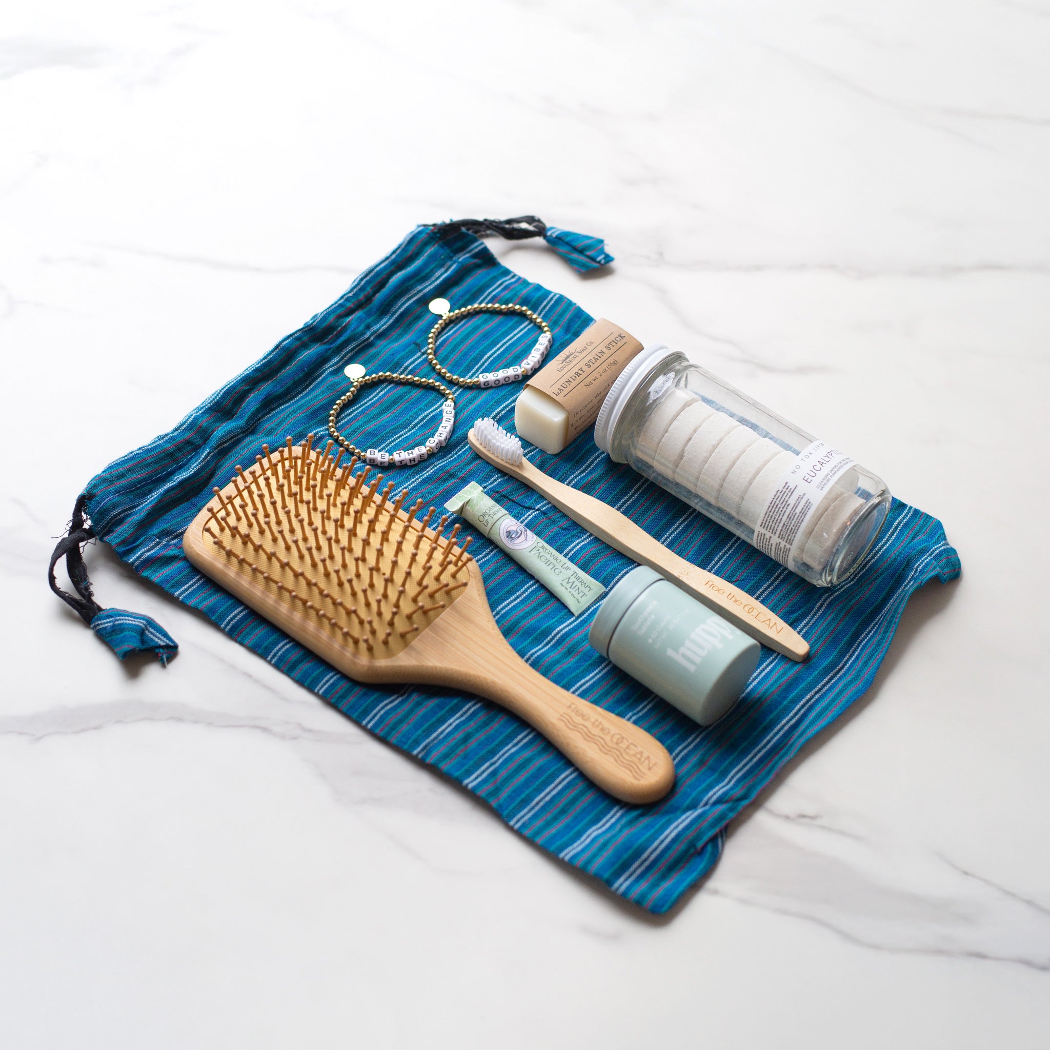 Eco-friendly self-care items arranged on a blue striped recycled drawstring bag, including a bamboo brush, toothbrush, toothpaste tabs, and more.
