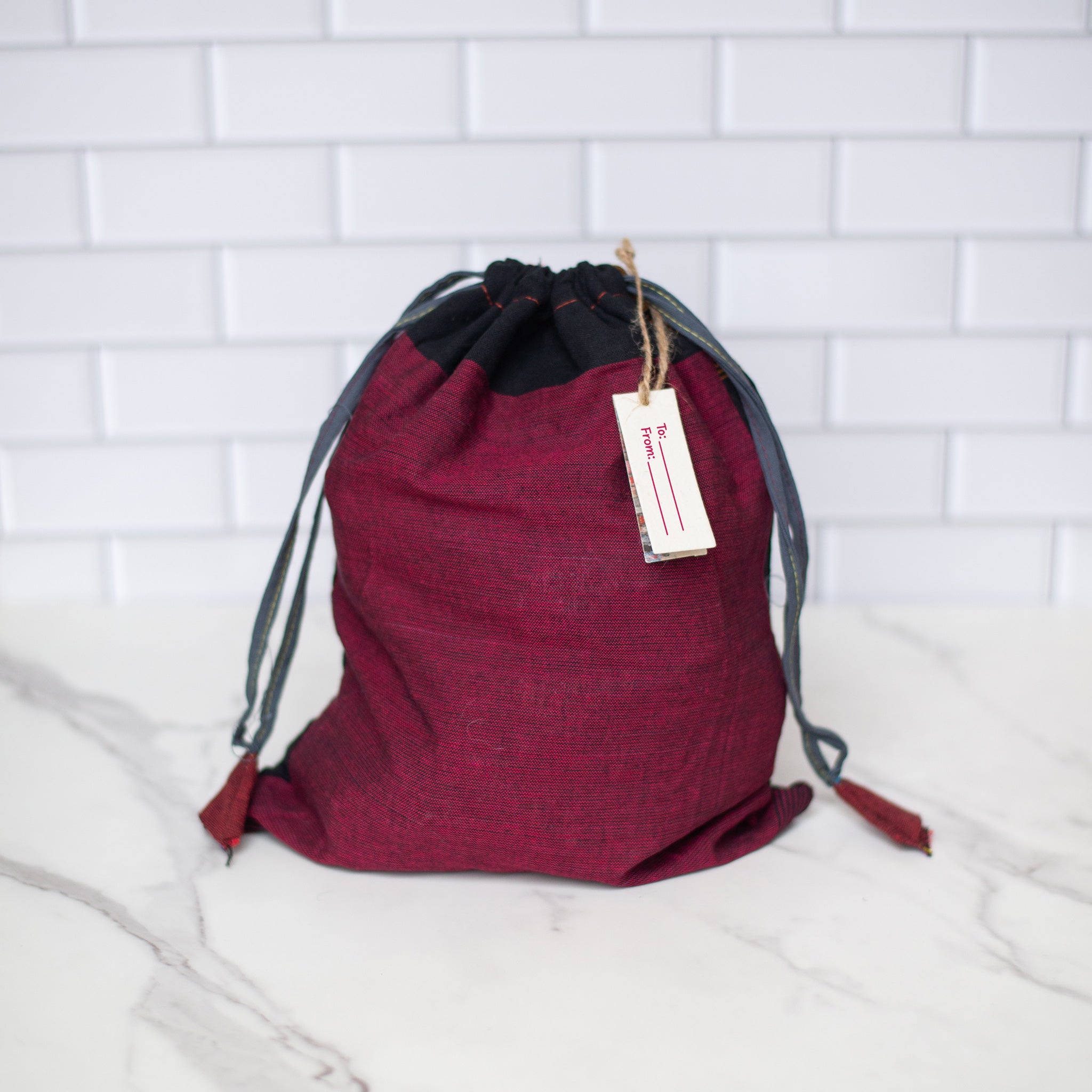 A red and black recycled drawstring bag with a gift tag attached, designed for reusable, eco-friendly gifting and everyday storage.