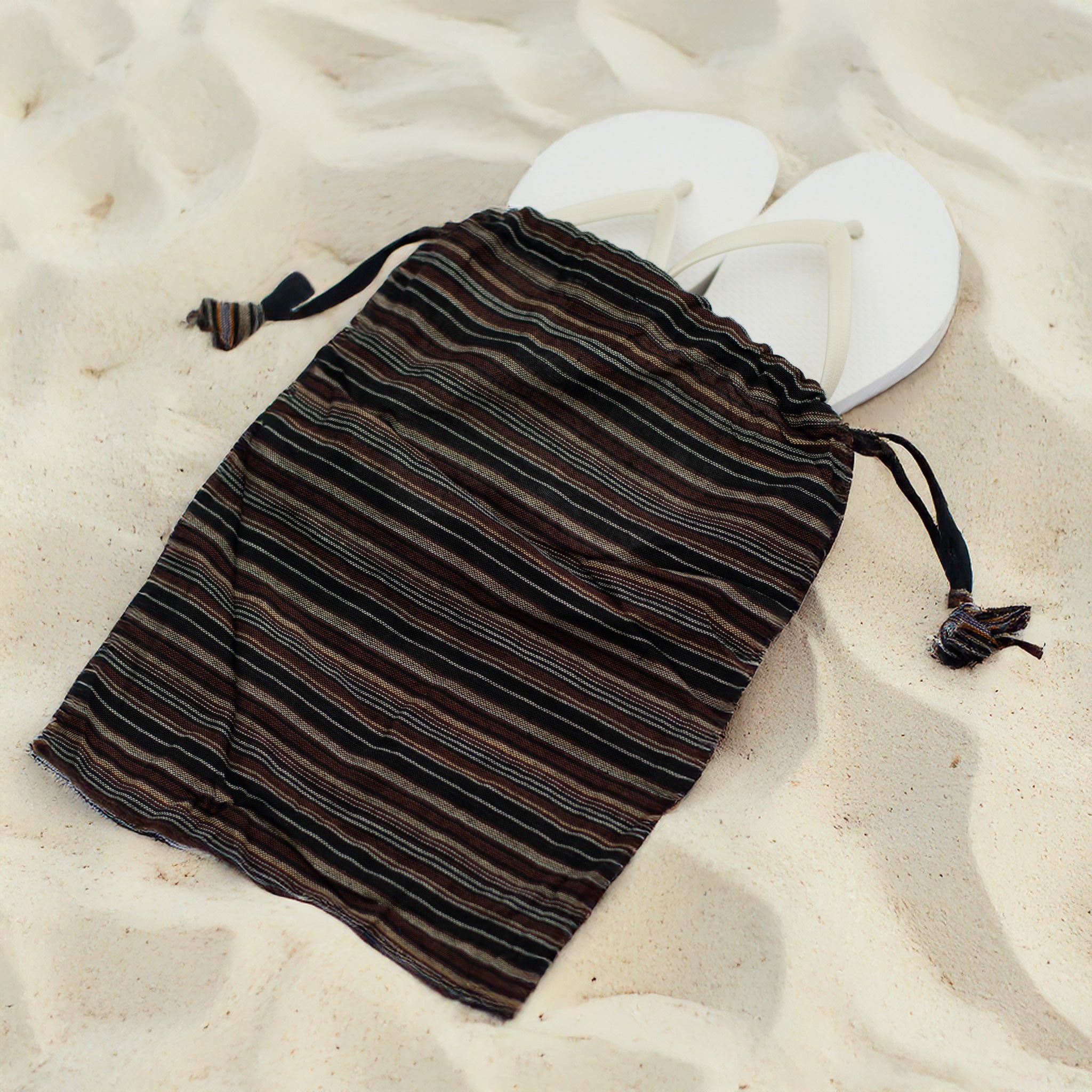 A brown and black striped recycled drawstring bag sits on sand with white flip-flops inside, showing its use as an eco-friendly beach essential.