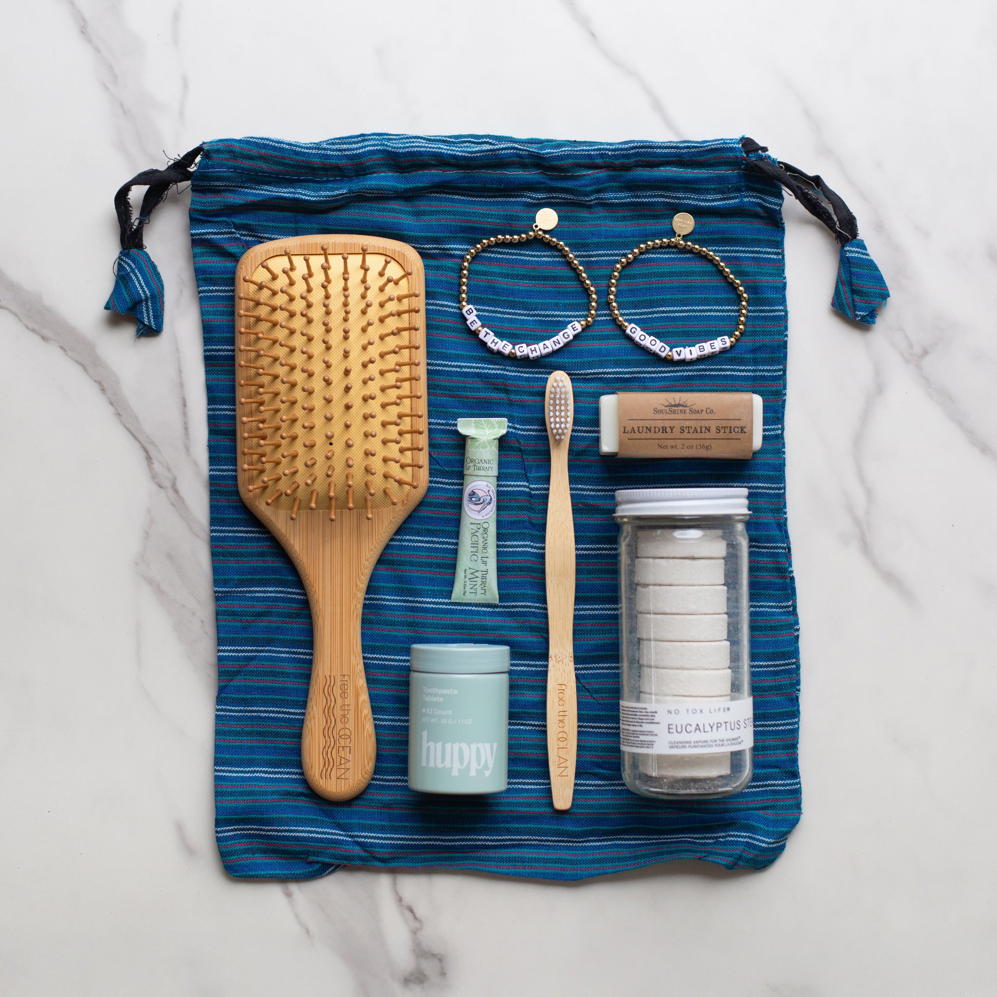 A blue recycled drawstring bag displayed with eco-friendly items like a bamboo brush, toothbrush, toothpaste tablets, and natural self-care products.