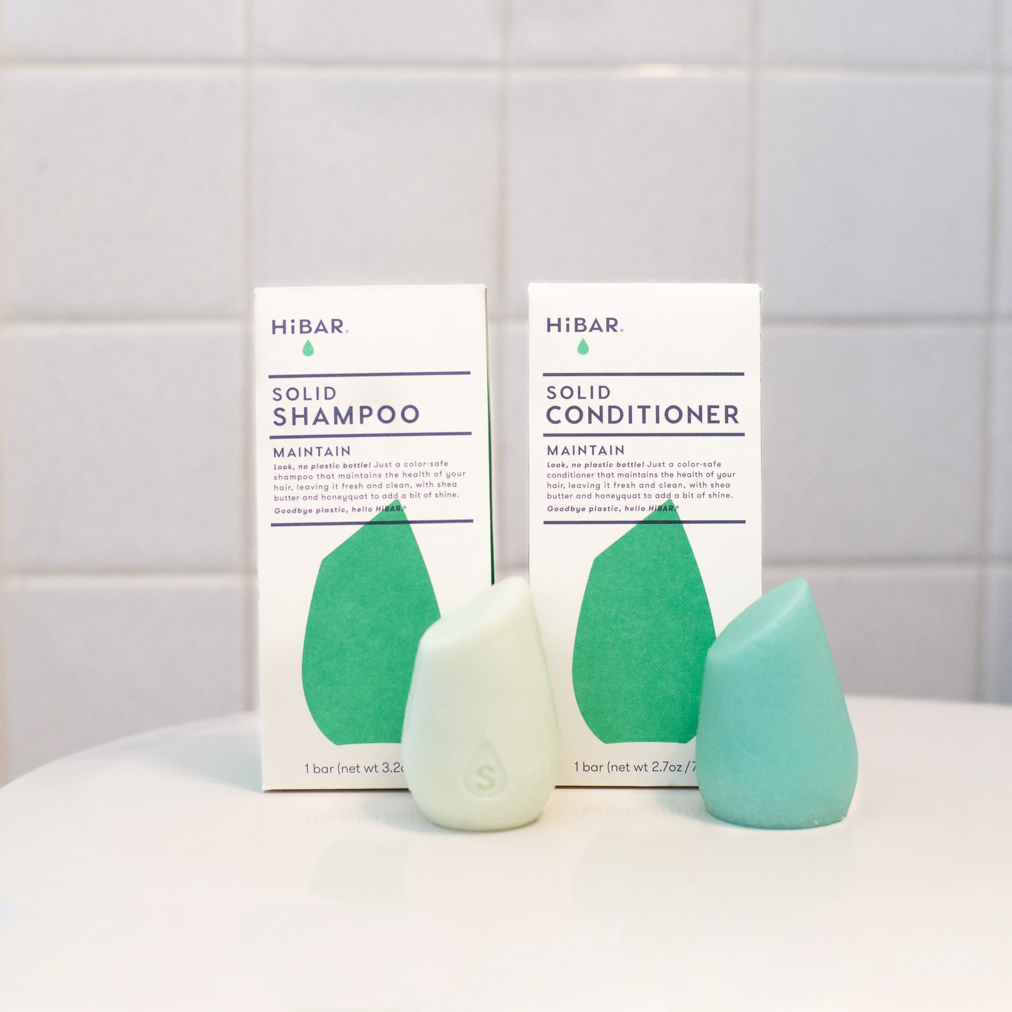 Two HiBar MAINTAIN solid hair care products: a light green shampoo bar and a light blue-green conditioner bar, featuring no plastic and made with organic ingredients like shea butter and honeyquat.